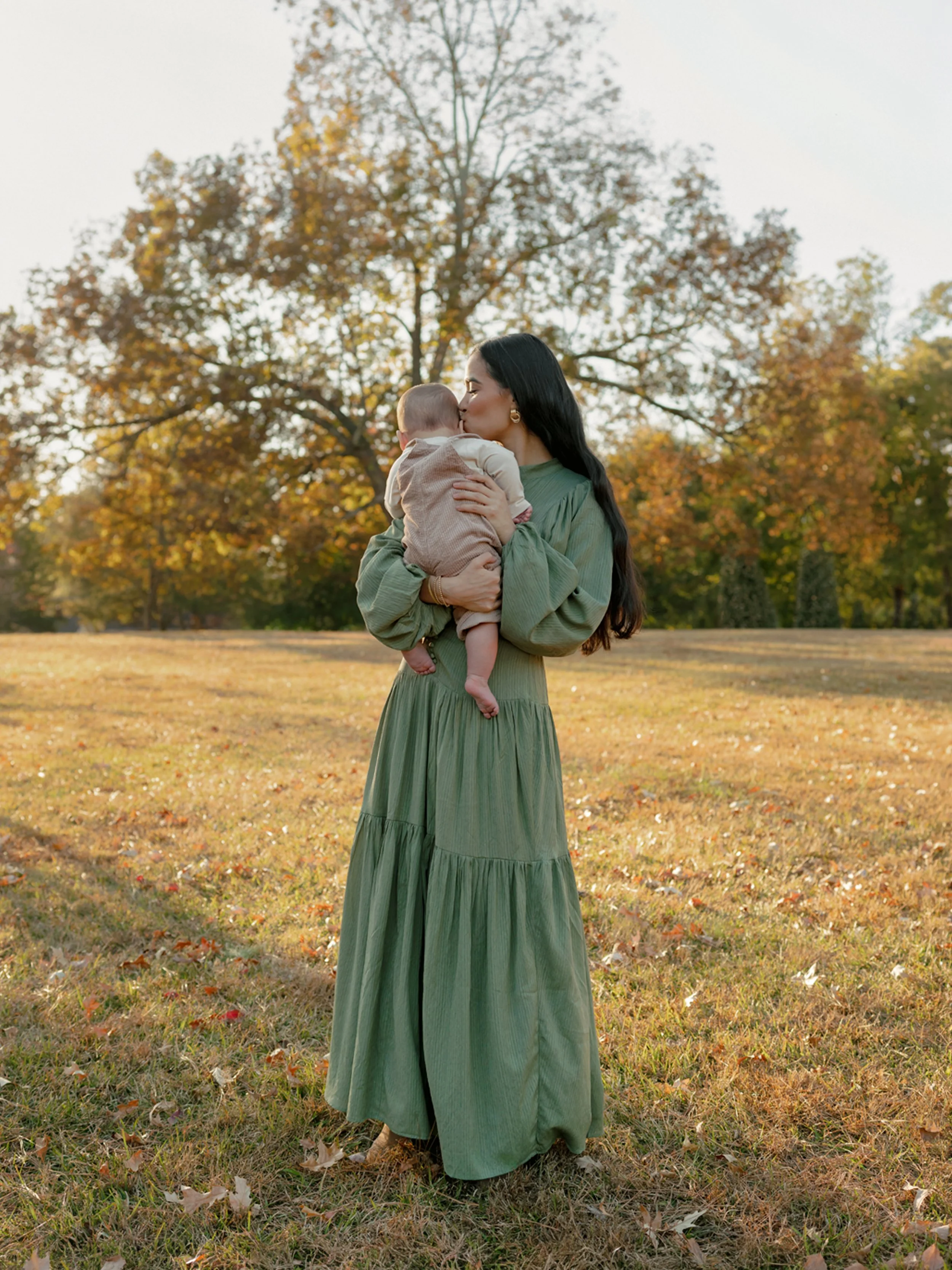 A woman in a green dress holding a baby in an outdoor park during fall, kissing the baby's forehead with trees displaying autumn leaves in the background.