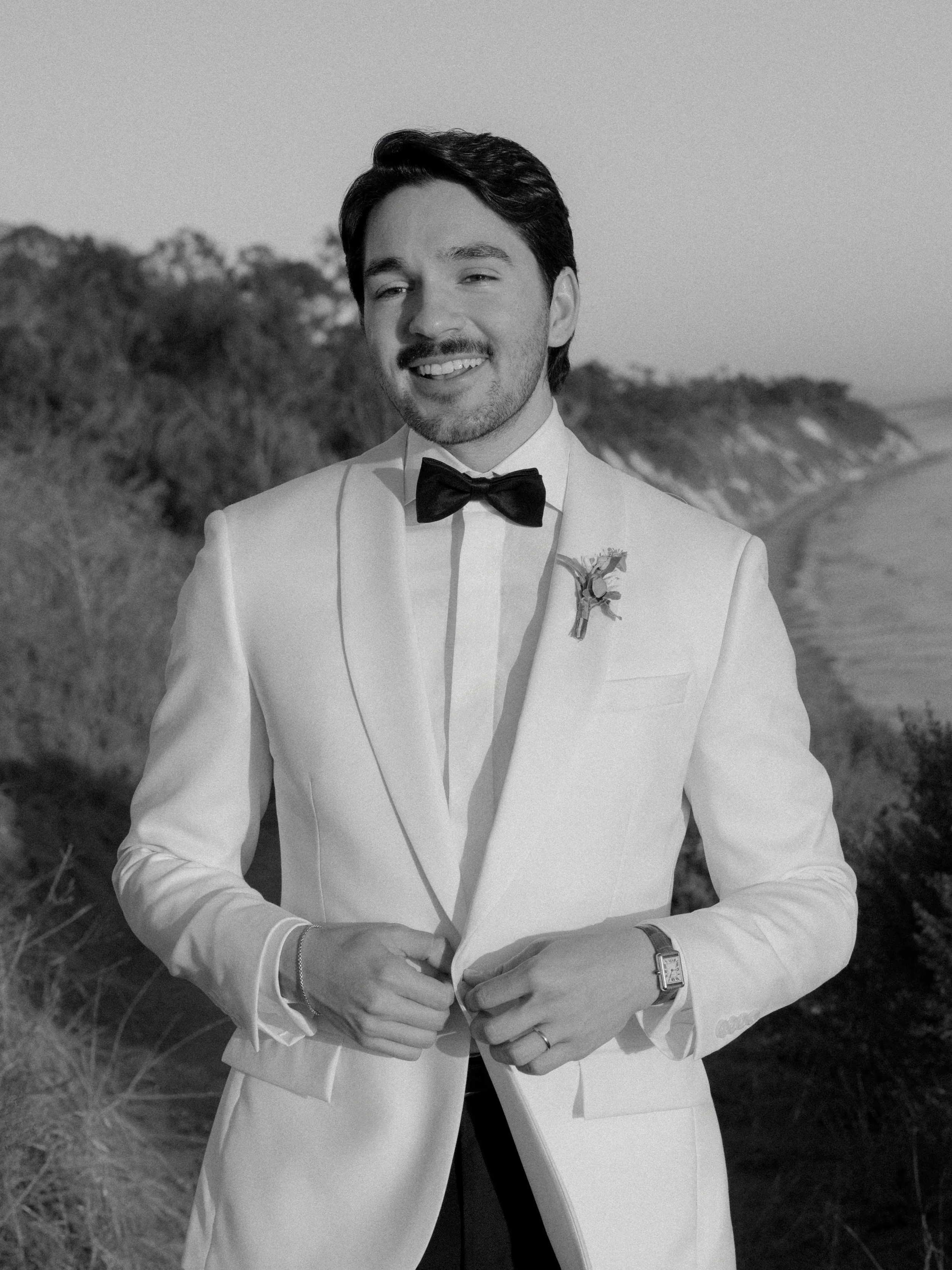 A young man with dark hair, dressed in a white tuxedo with a black bow tie, standing outdoors on a beach or coastal area with trees in the background, smiling at the camera.