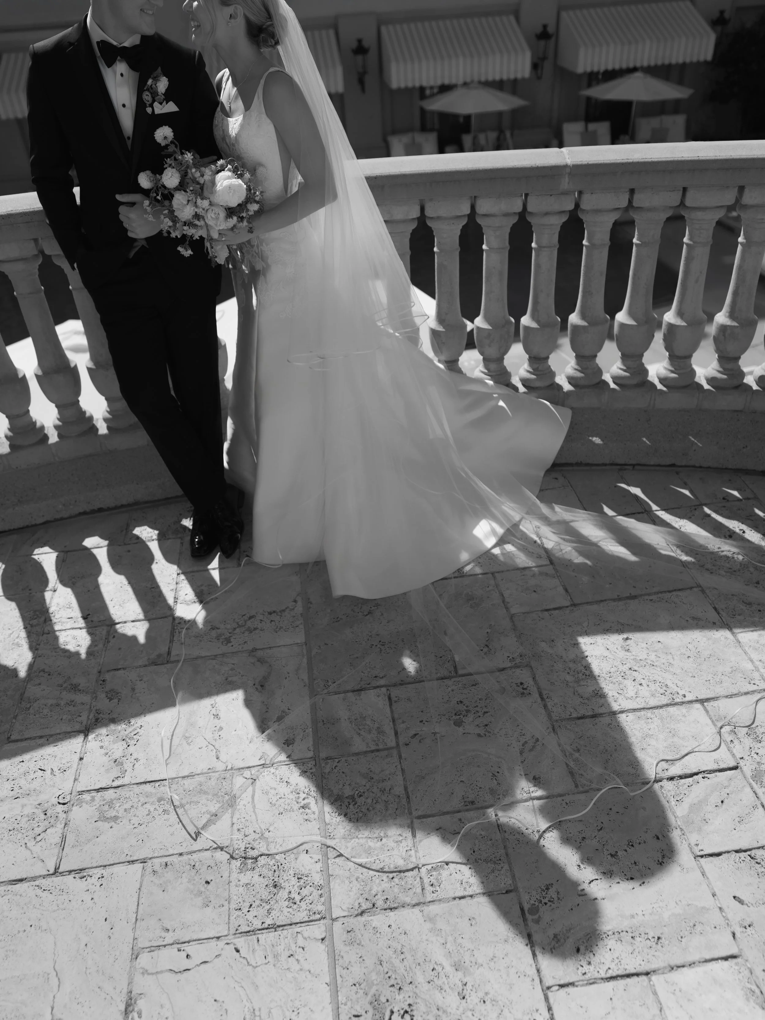 Black and white photo of a bride and groom on a balcony, smiling and leaning close. The bride holds a bouquet and wears a wedding gown with a long veil, while the groom is dressed in a tuxedo. Shadows cast on the tiled floor beneath them.