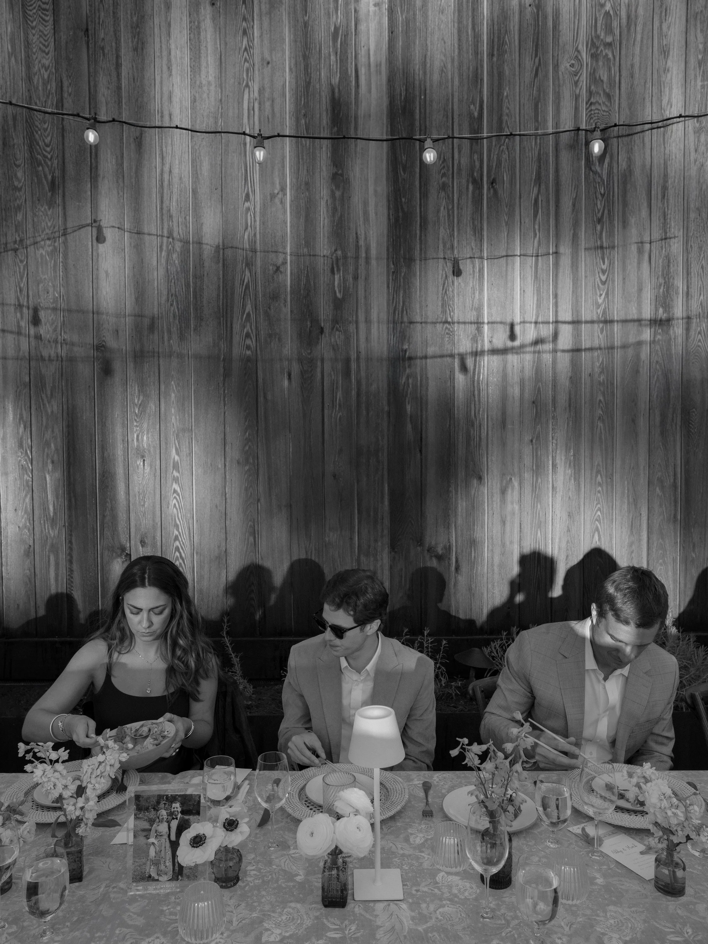People sitting at a table during a dinner event, with a wooden wall and string lights in the background.