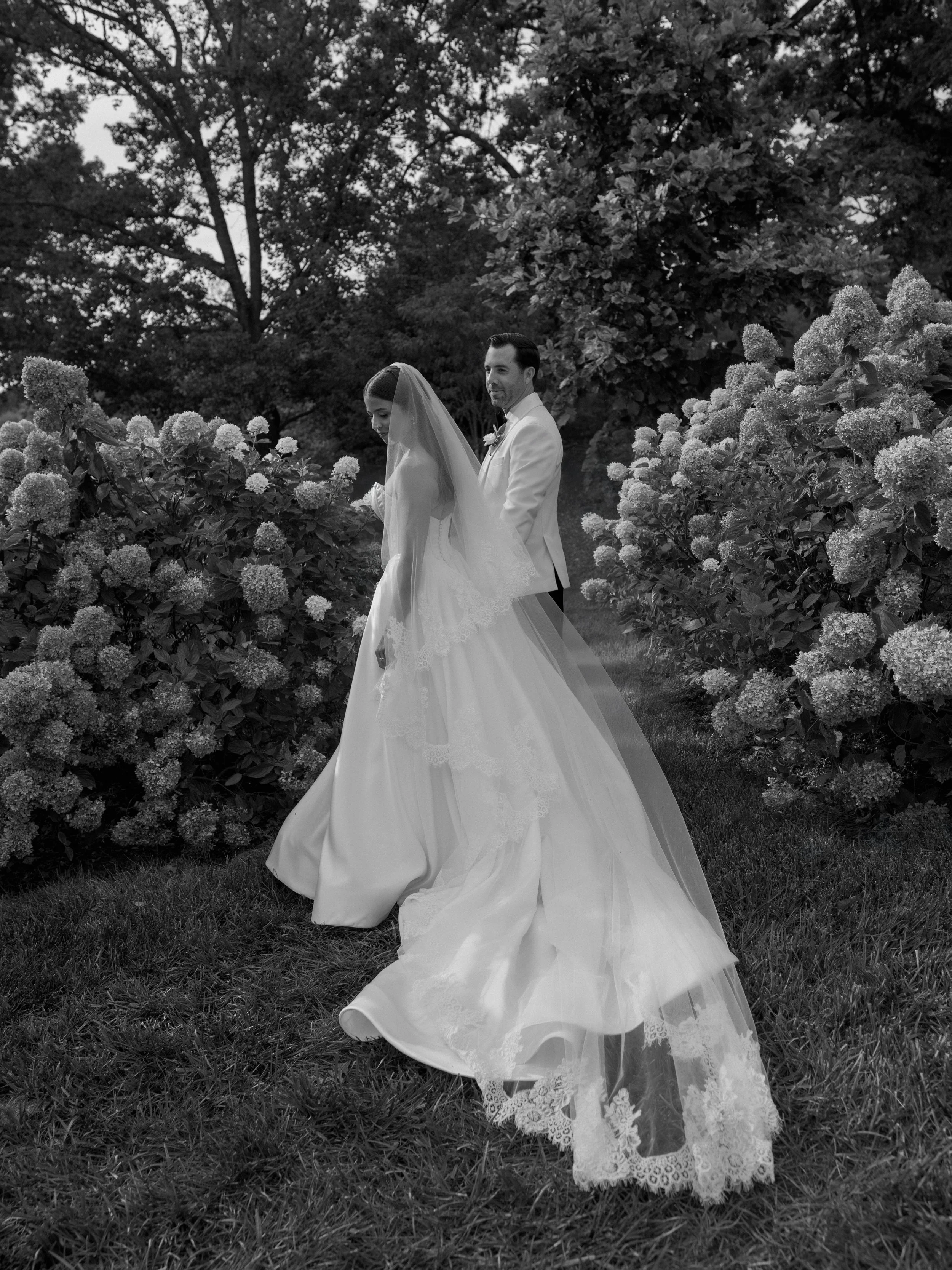 A black-and-white photo of a bride and groom outdoors among bushes and trees. The bride is wearing a long wedding dress with a veil, and the groom is wearing a suit. The couple is walking along a path, with the groom looking towards the bride.