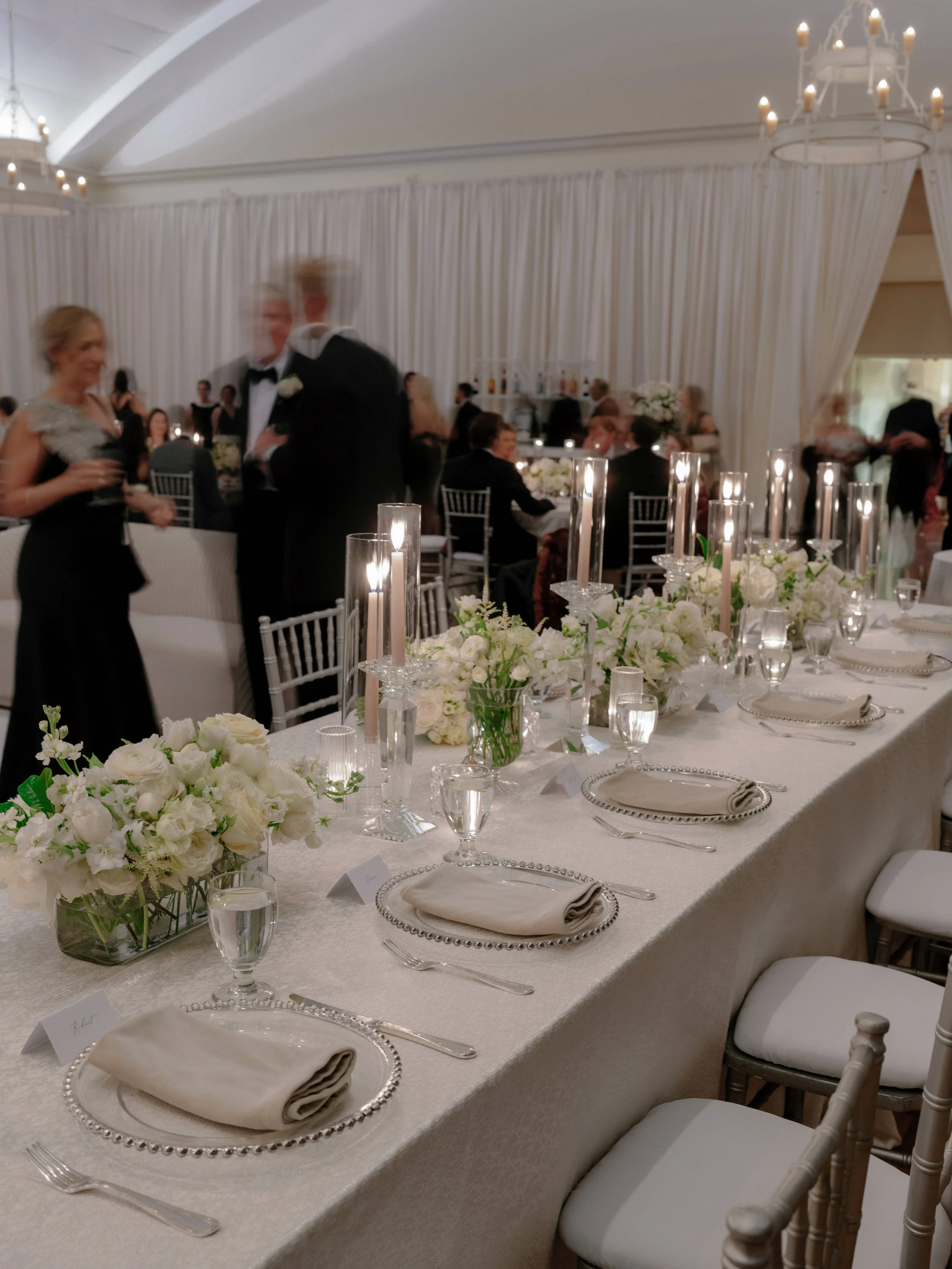 A decorated banquet table at a formal event with white floral centerpieces, tall candles in glass holders, place settings with napkins, silverware, and clear water glasses, with blurred people in tuxedos and elegant dresses in the background.