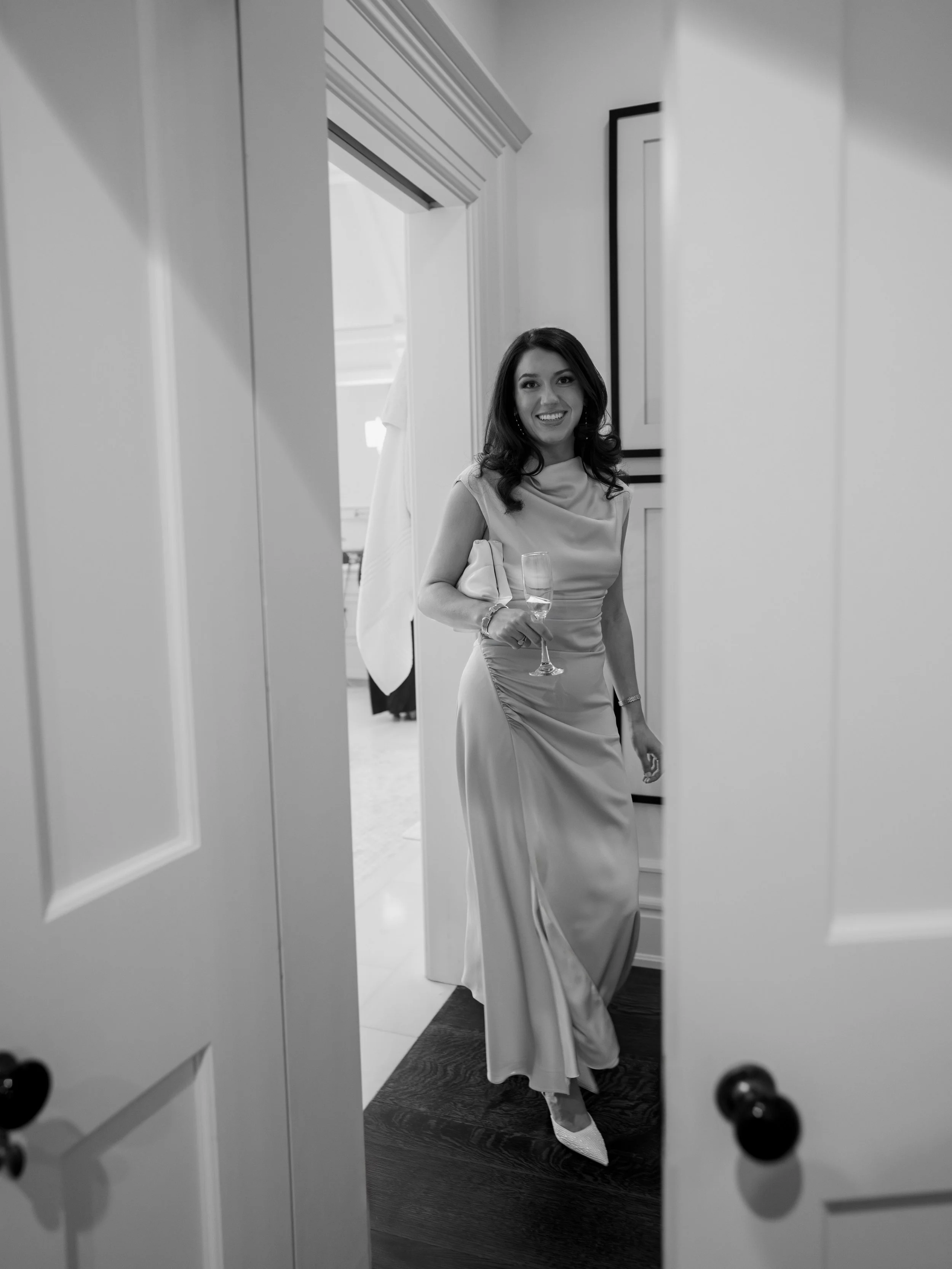A woman in an elegant dress holding a glass of champagne, smiling, as she stands in a doorway.