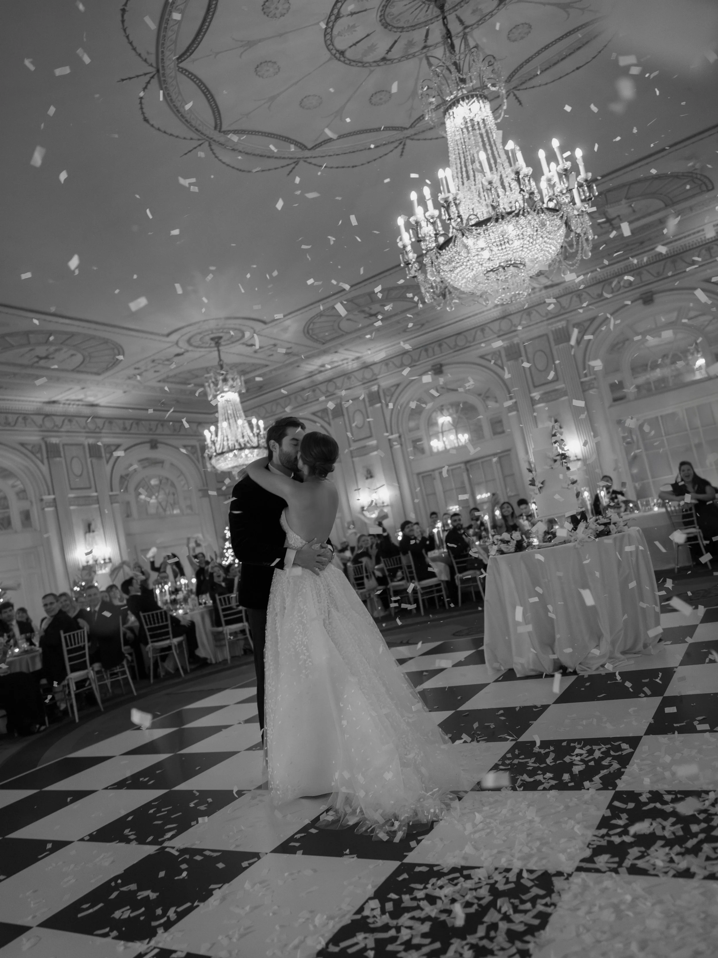 A newlywed couple sharing their first dance at a wedding reception in a grand ballroom decorated with chandeliers and confetti falling from the ceiling.