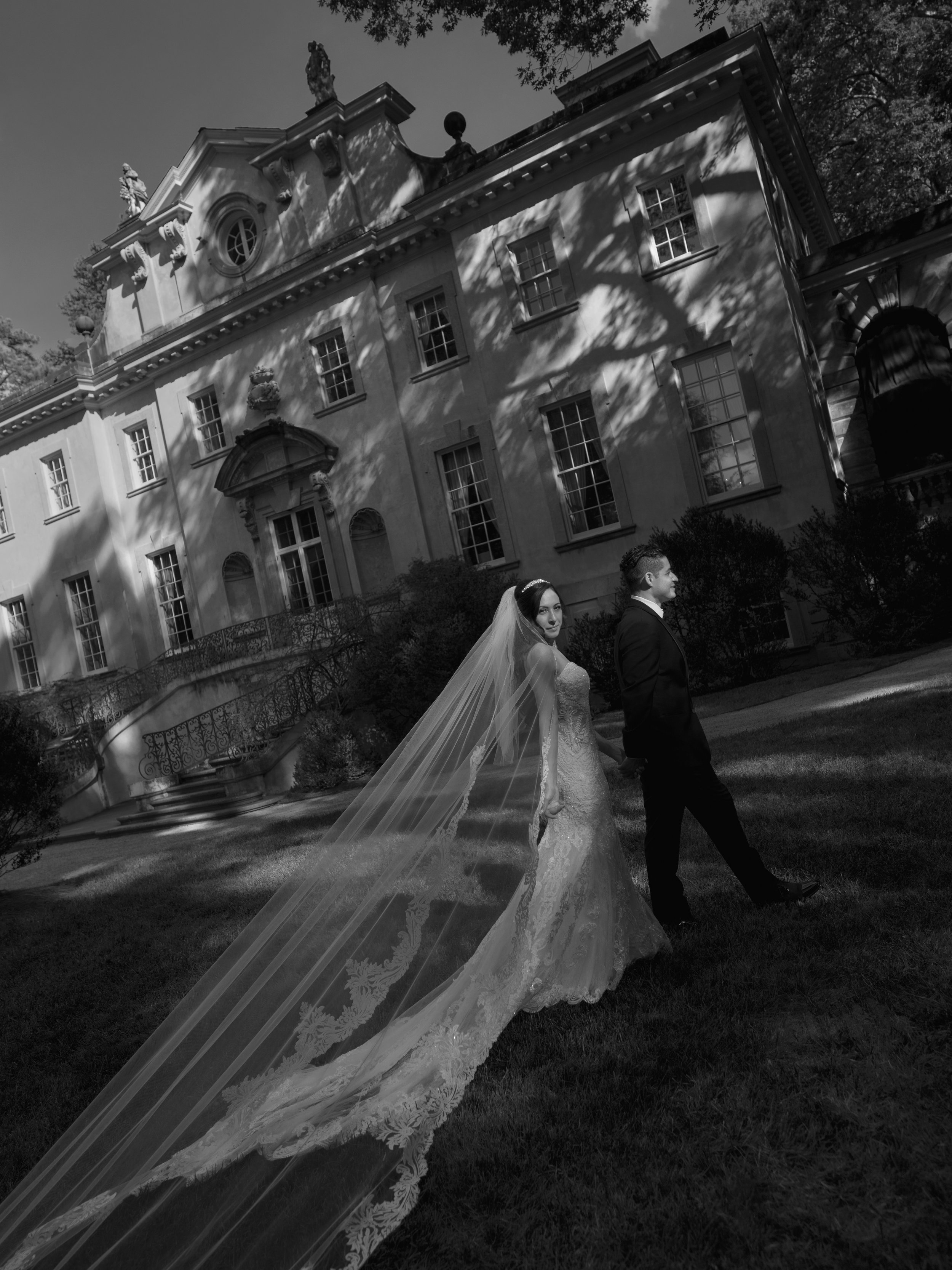 Black and white photograph of a bride and groom walking outside a large, historic mansion with multiple windows and ornate architecture. The bride is wearing a lace wedding gown and a long veil, and the groom is in a tuxedo. Shadows of trees are cast