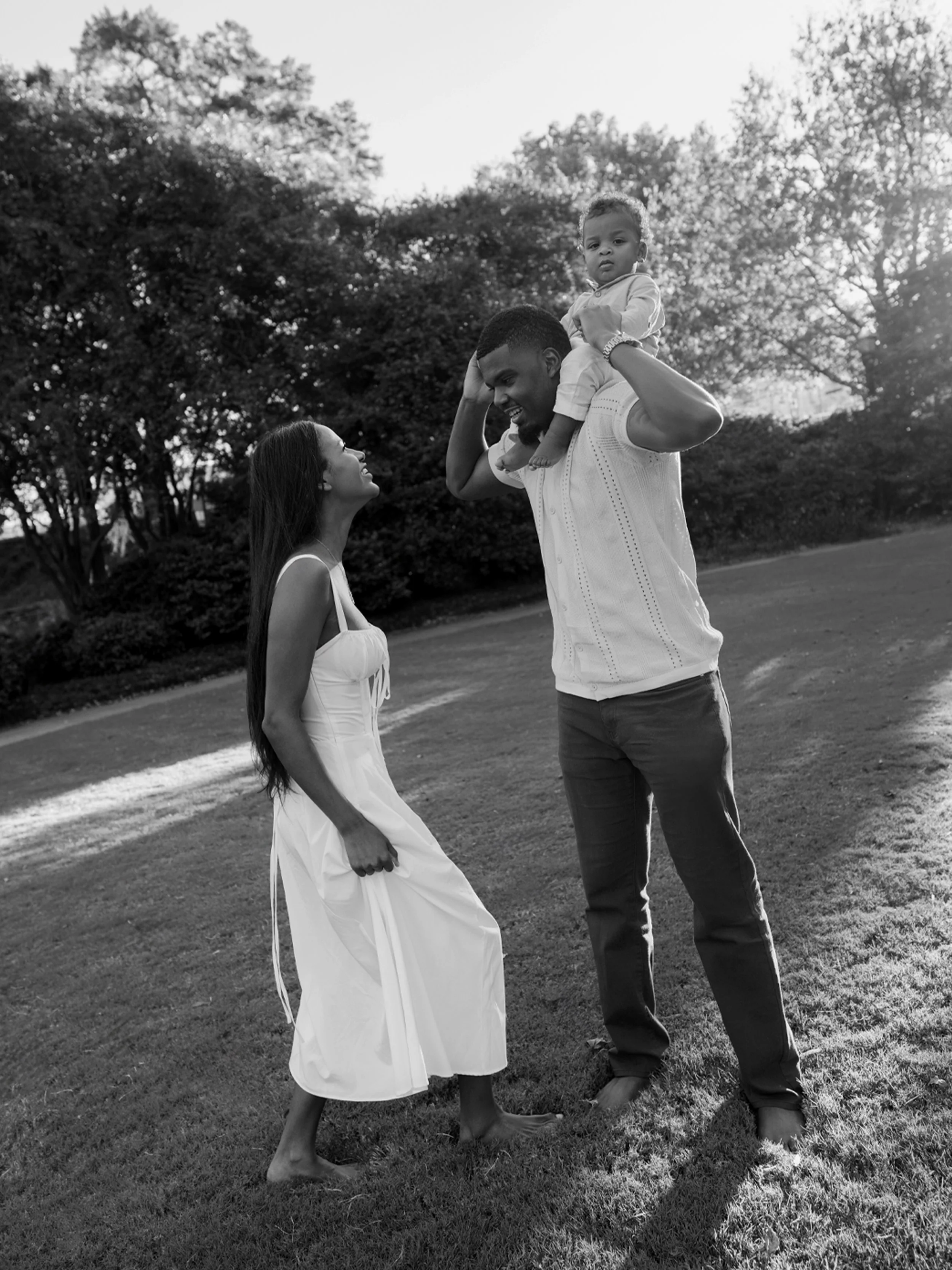 A black and white photo of a family in a park with trees in the background. A man is holding a young child on his shoulders, and a woman is standing in front of them, they are looking at each other and smiling.