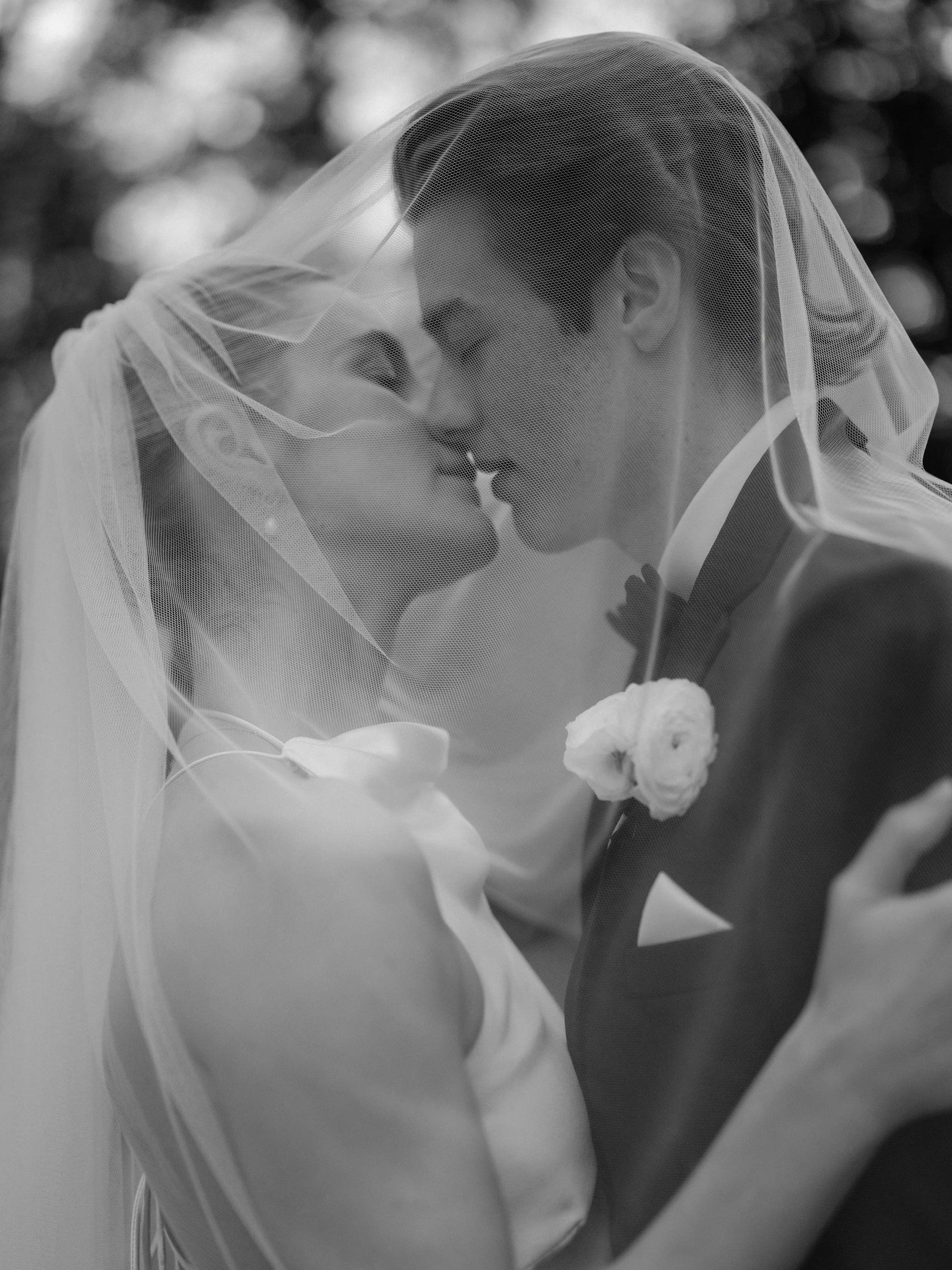 Black and white photo of a bride and groom sharing a kiss under a veil during their wedding.