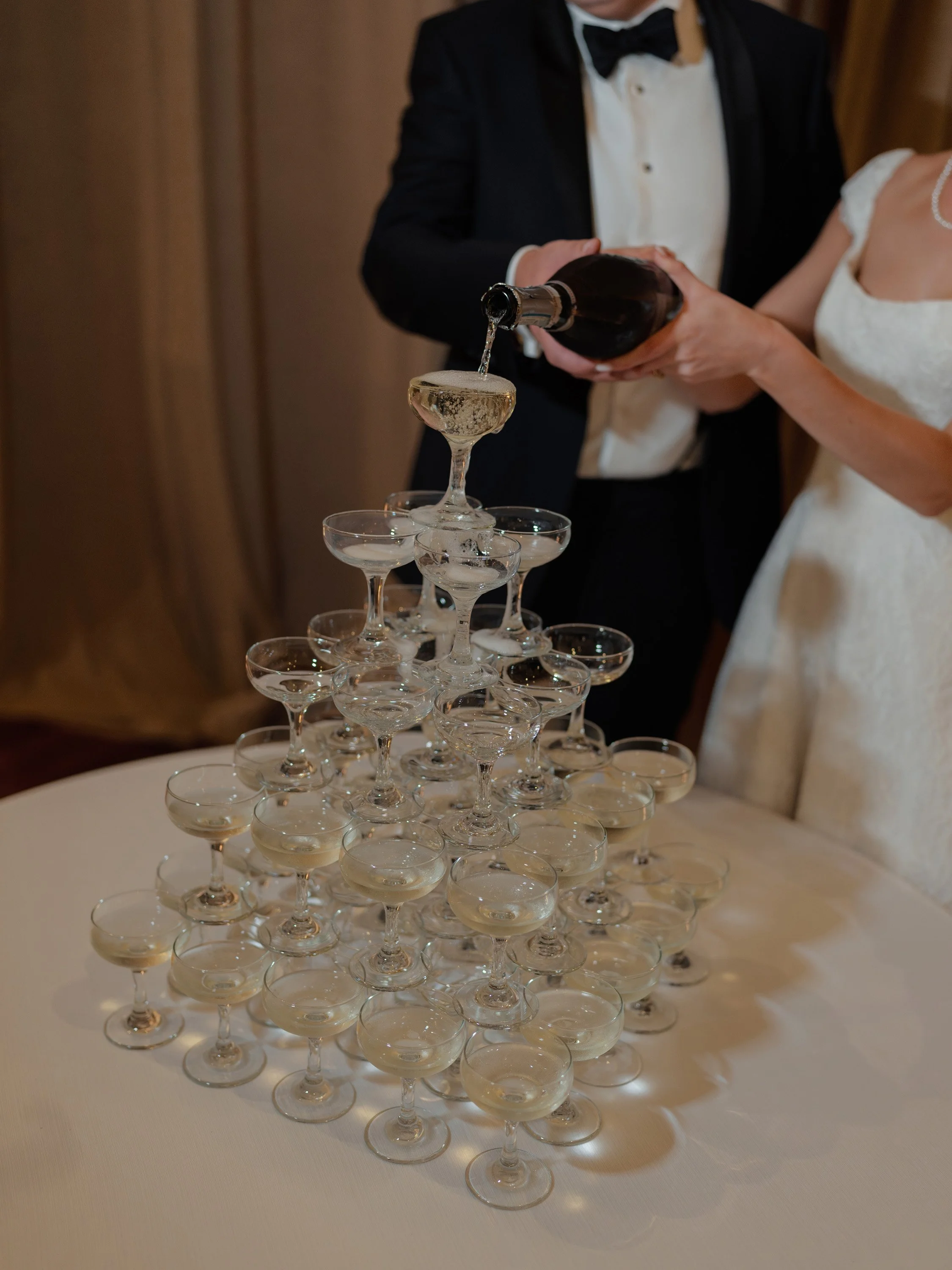A person in a tuxedo and a person in a wedding dress are pouring champagne into a tower of glasses.