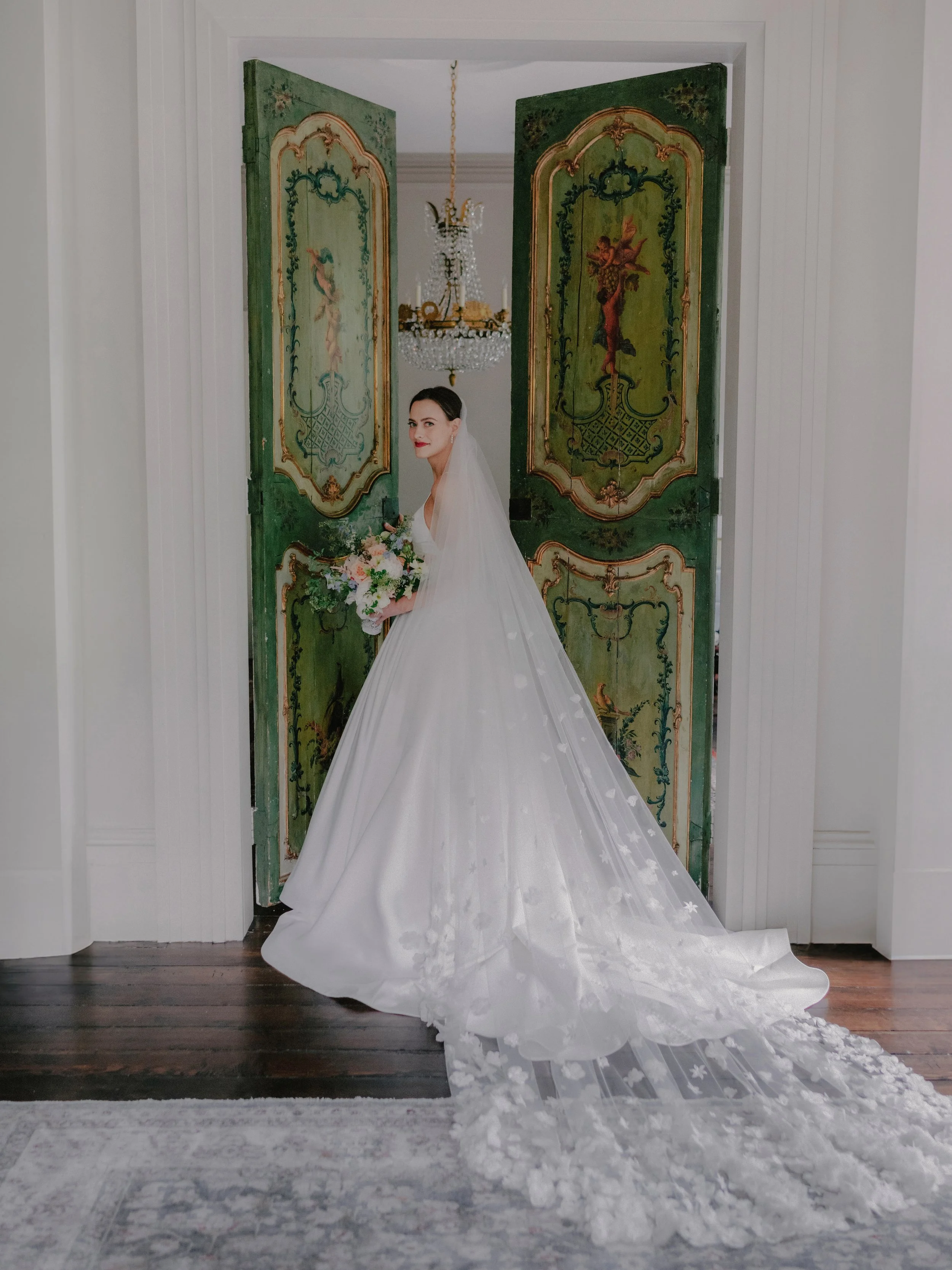 A bride in a white wedding gown holding a bouquet stands in front of ornate green double doors with vintage artwork, with a chandelier hanging overhead.