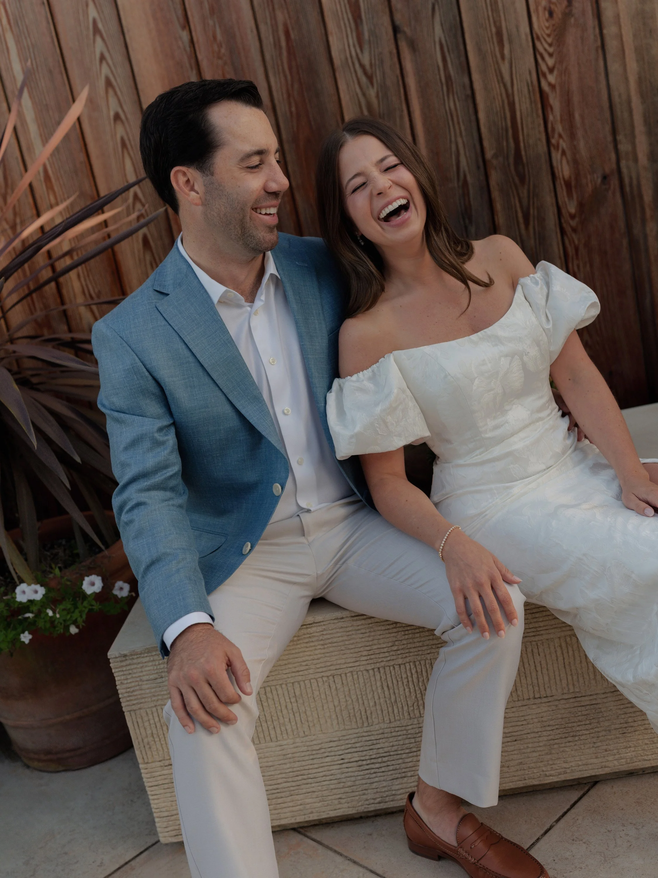 A man in a light blue blazer and cream pants sitting next to a woman in a white off-shoulder dress, both laughing and enjoying a moment together outdoors with wooden fencing and plants in the background.