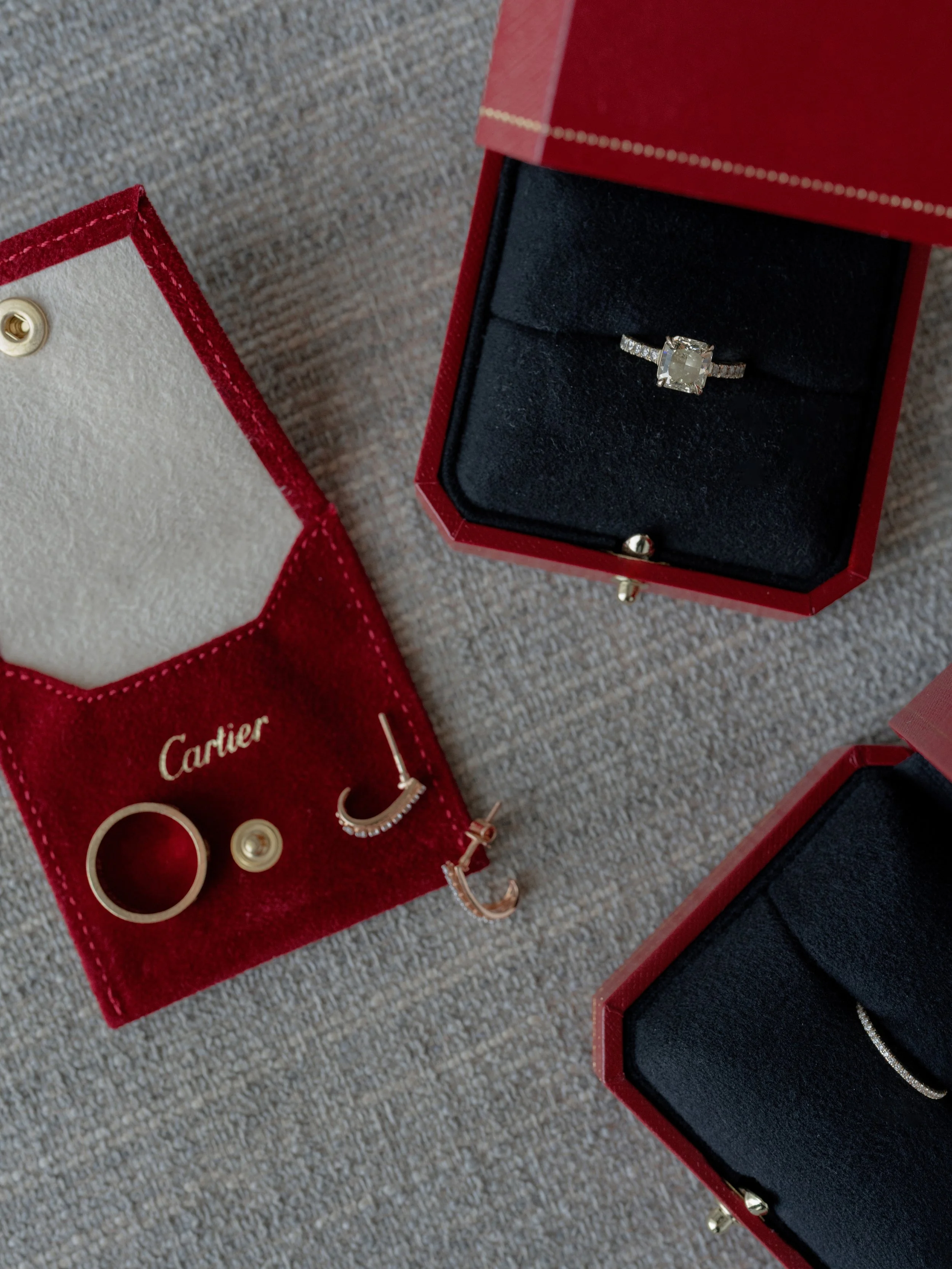 A diamond ring in a red Cartier jewelry box, jewelry box with black interior, and a pair of rose gold hoop earrings with diamonds, all on a textured surface.