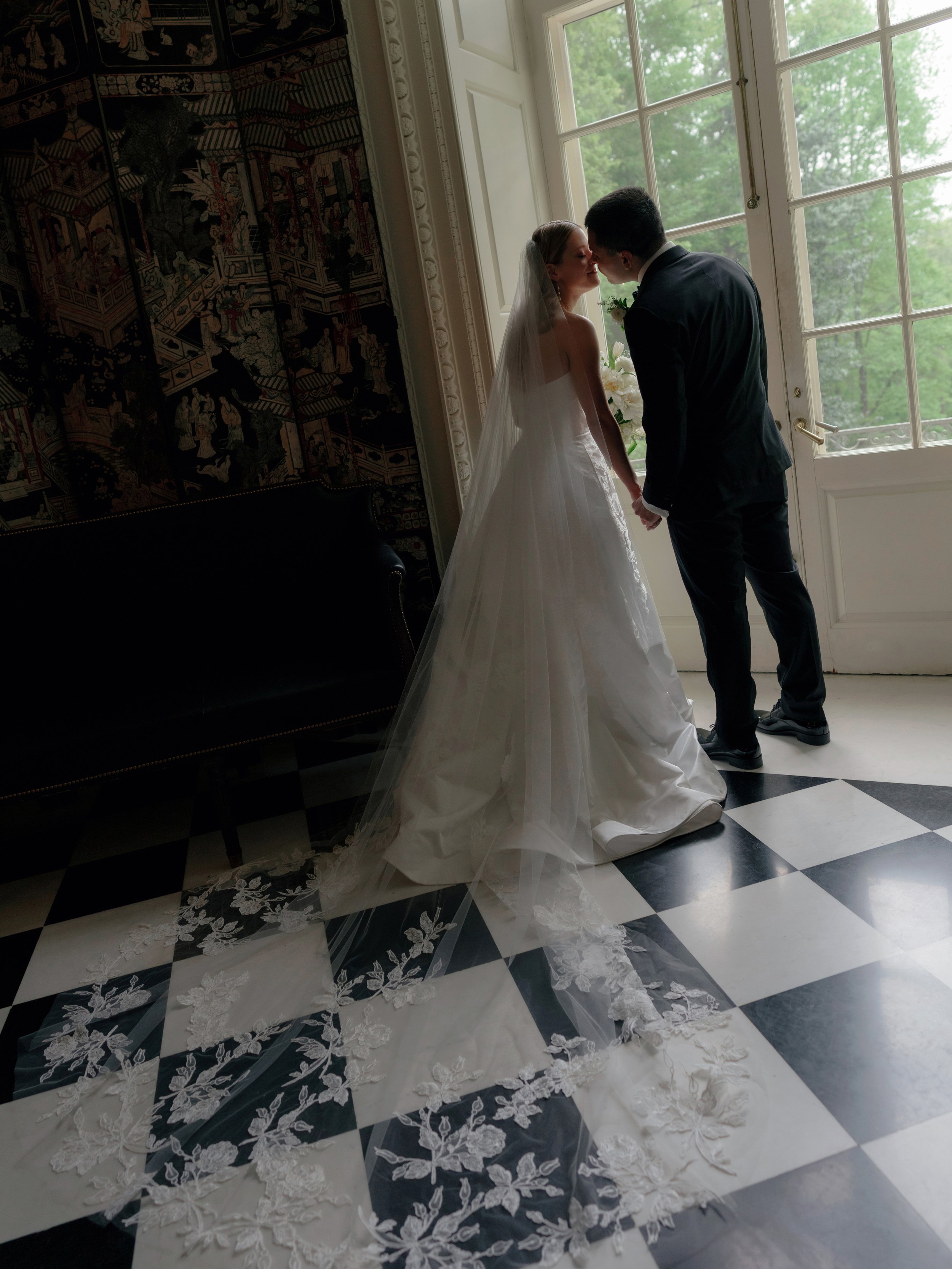 A bride and groom stand close together in front of large windows, holding hands and leaning in for a kiss. The bride wears a white wedding gown with a long lace train and veil, while the groom wears a black suit.