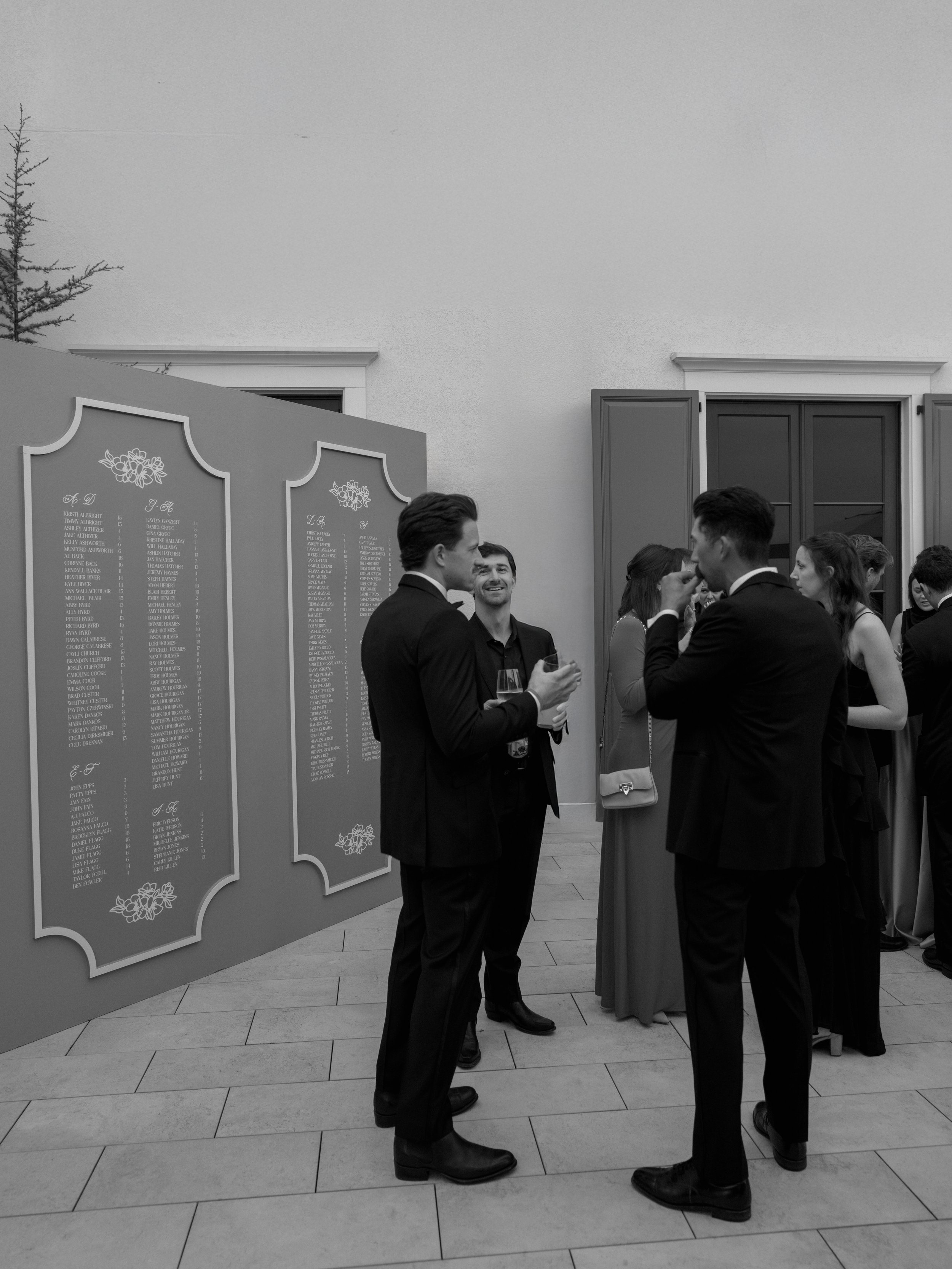 Group of men and women dressed in formal attire socializing at an event, with a large placard displaying a list of names in the background.