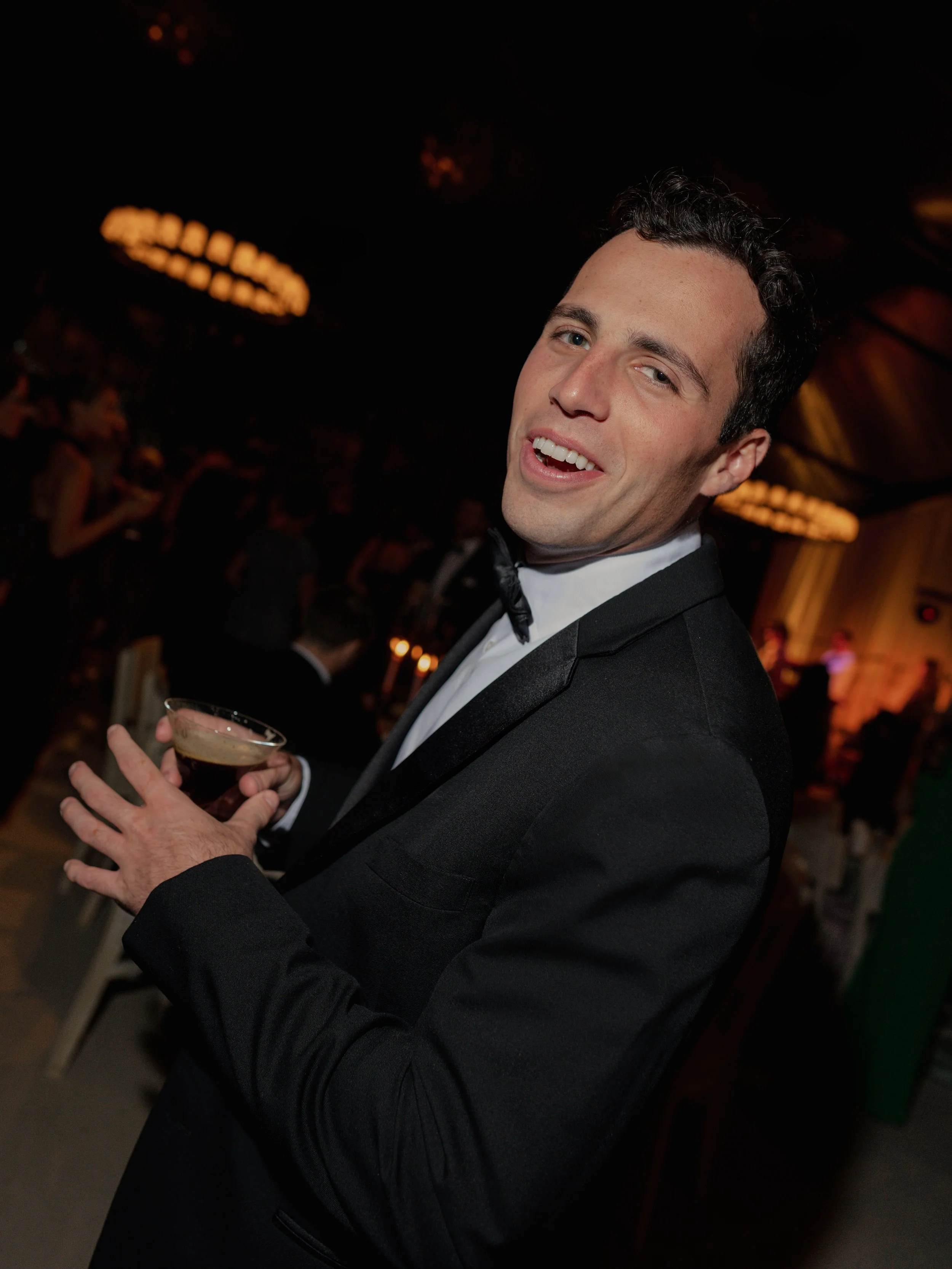 A man in a tuxedo holding a cocktail at a party or event.