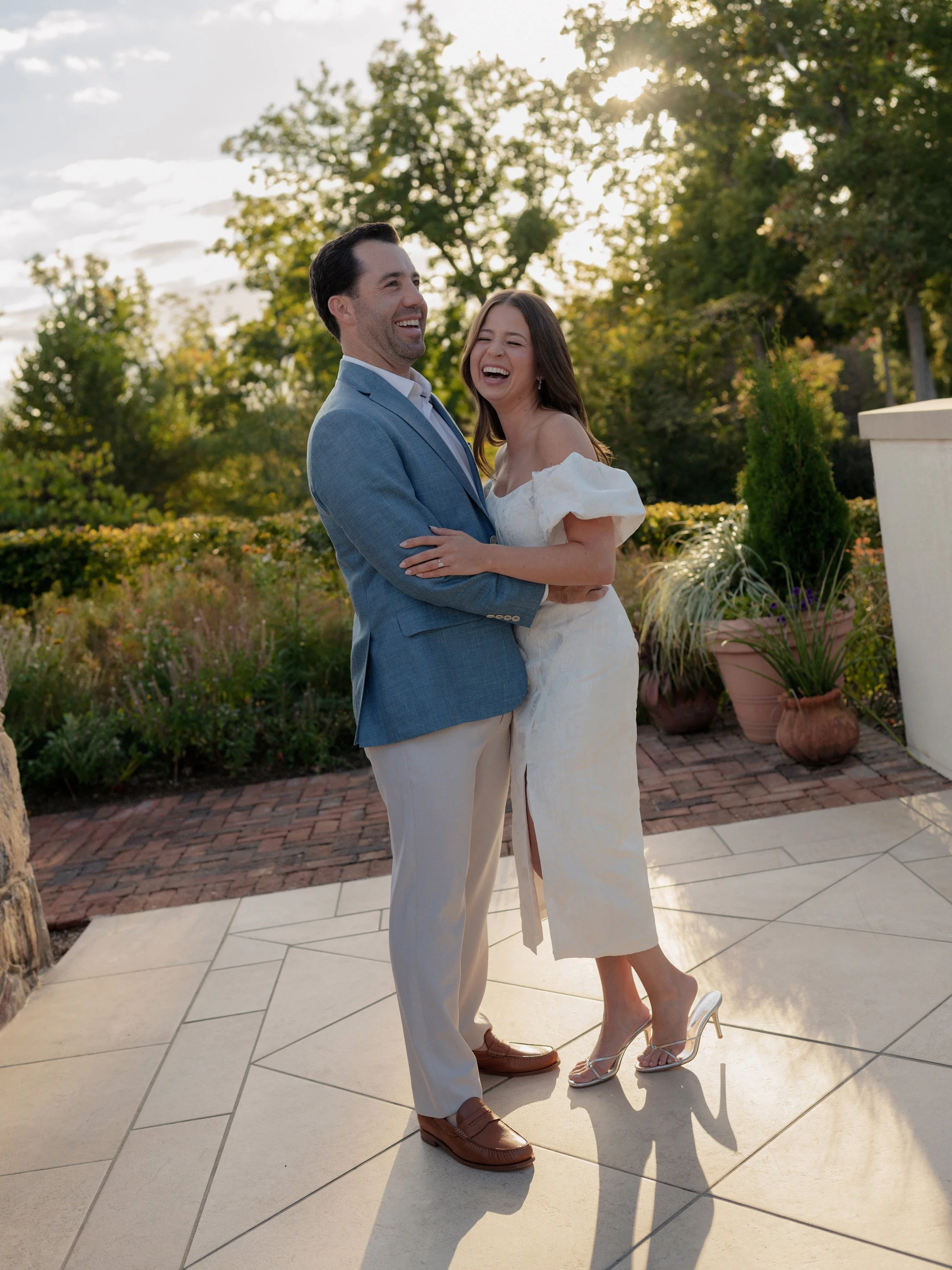 A couple laughing and hugging outdoors on a bright, sunny day, with greenery and potted plants in the background.