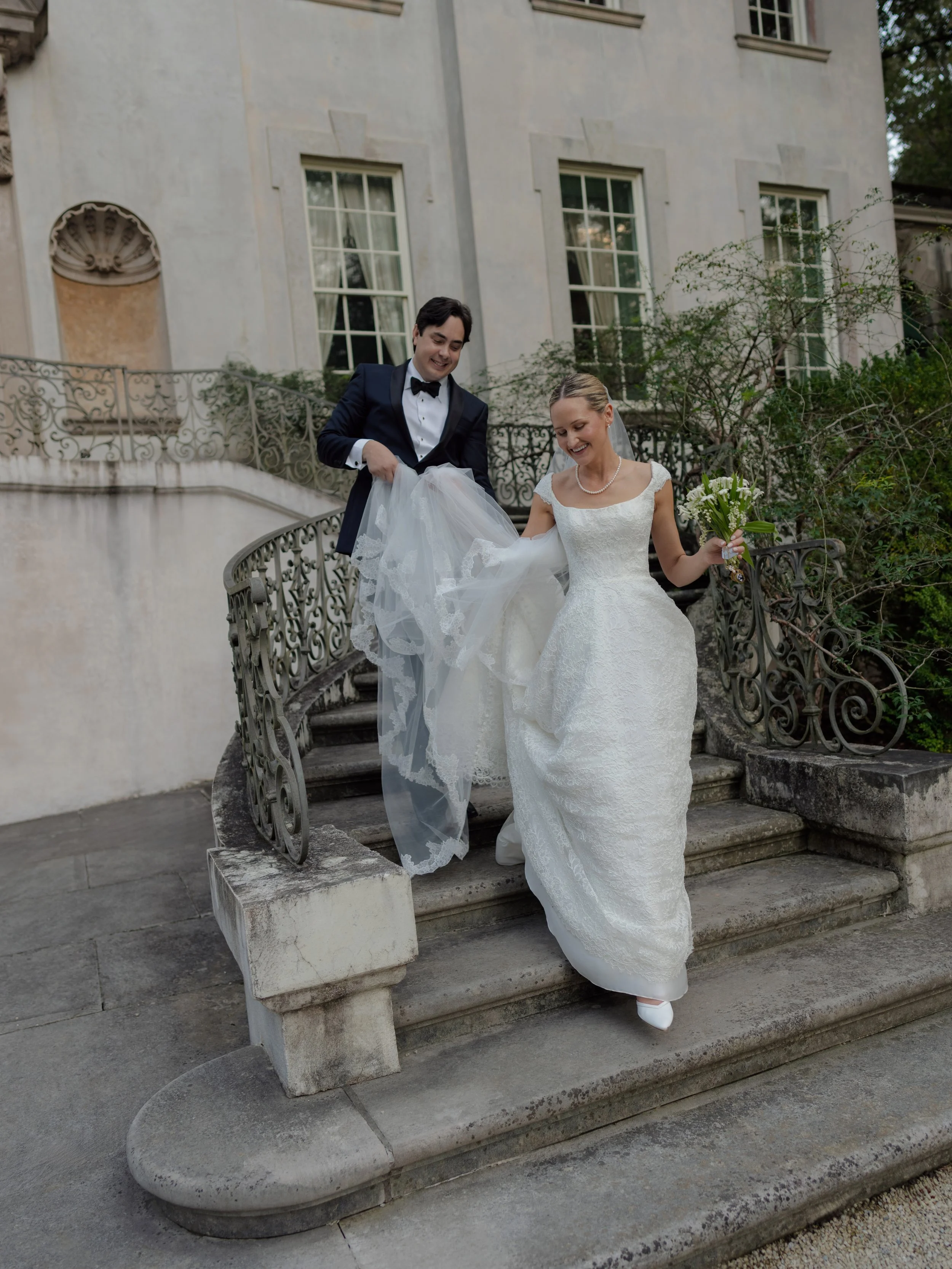 A bride and groom descending outdoor stone stairs, with the groom lifting the bride's dress, both smiling. The bride holds a bouquet of white flowers, and the groom wears a black tuxedo with a bow tie. The background shows a historic building with la