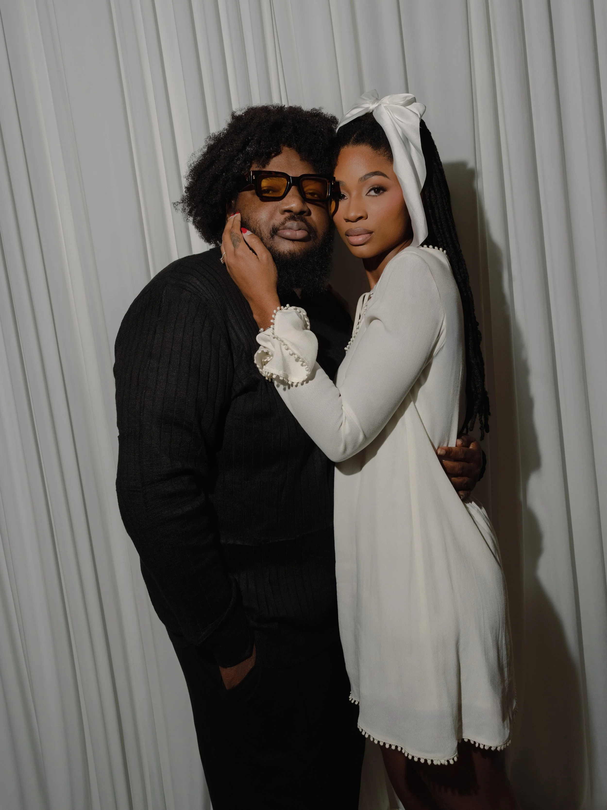 A man with glasses and a woman with long hair wearing a white headwrap, standing close together against a white curtain background, posing intimately for a photo.