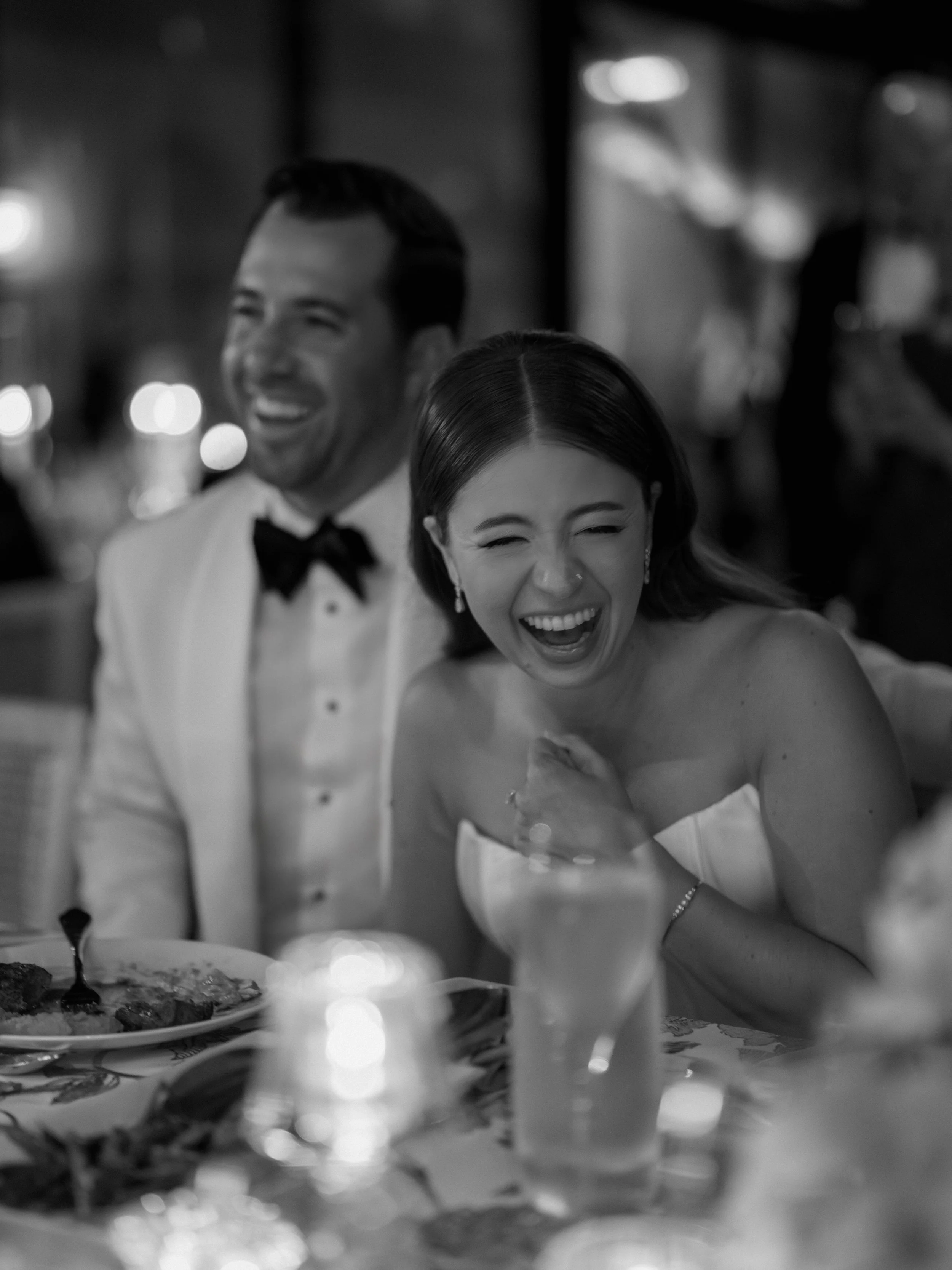 Black and white photo of a celebration with two people, a man in a tuxedo and a woman in a strapless dress, both laughing at a dinner table.