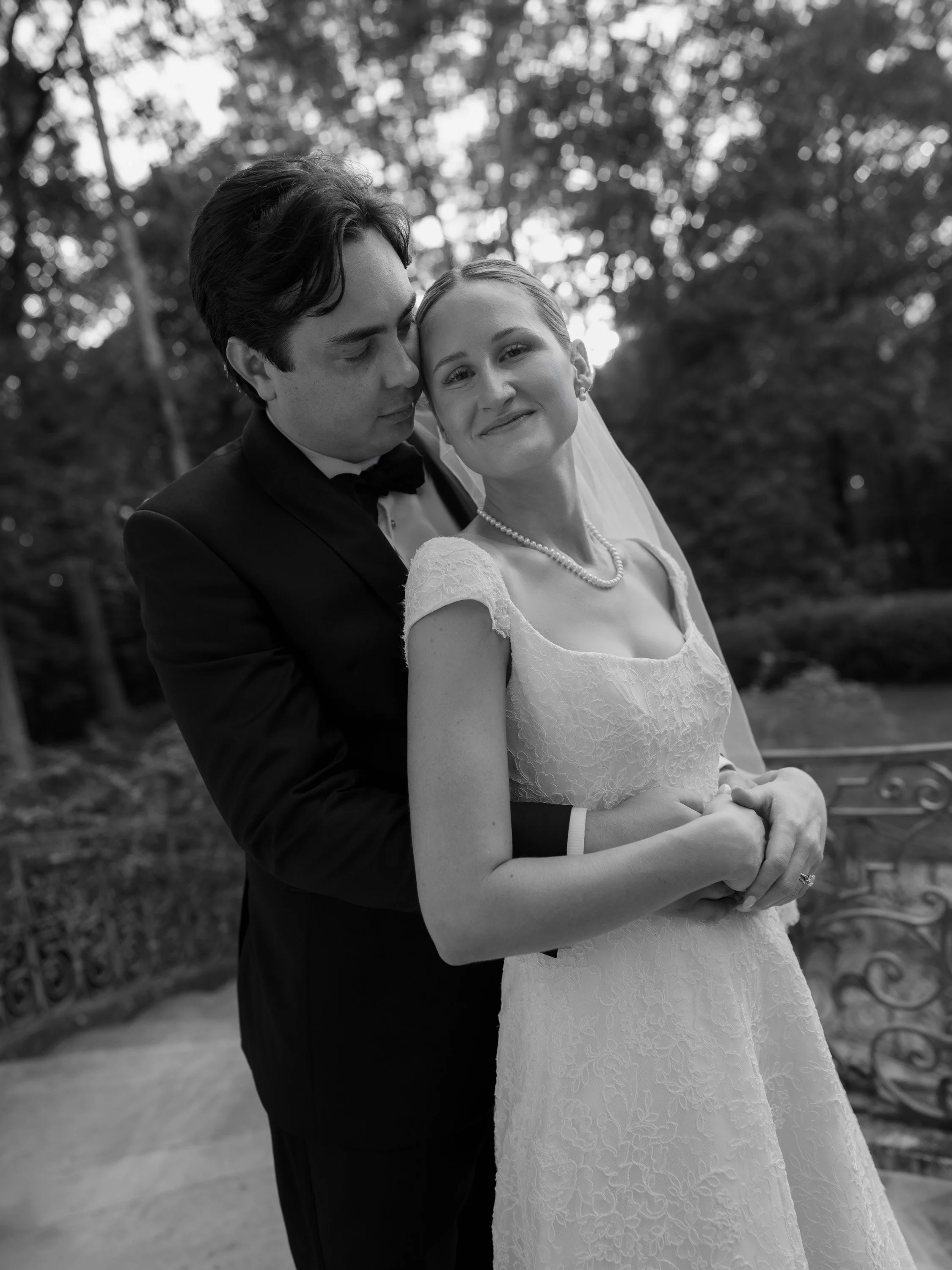 Black and white photo of a bride and groom embracing outdoors, with trees in the background. The bride is smiling with her head tilted slightly, wearing a lace wedding dress and pearls, while the groom is in a tuxedo and leaning towards her with clos