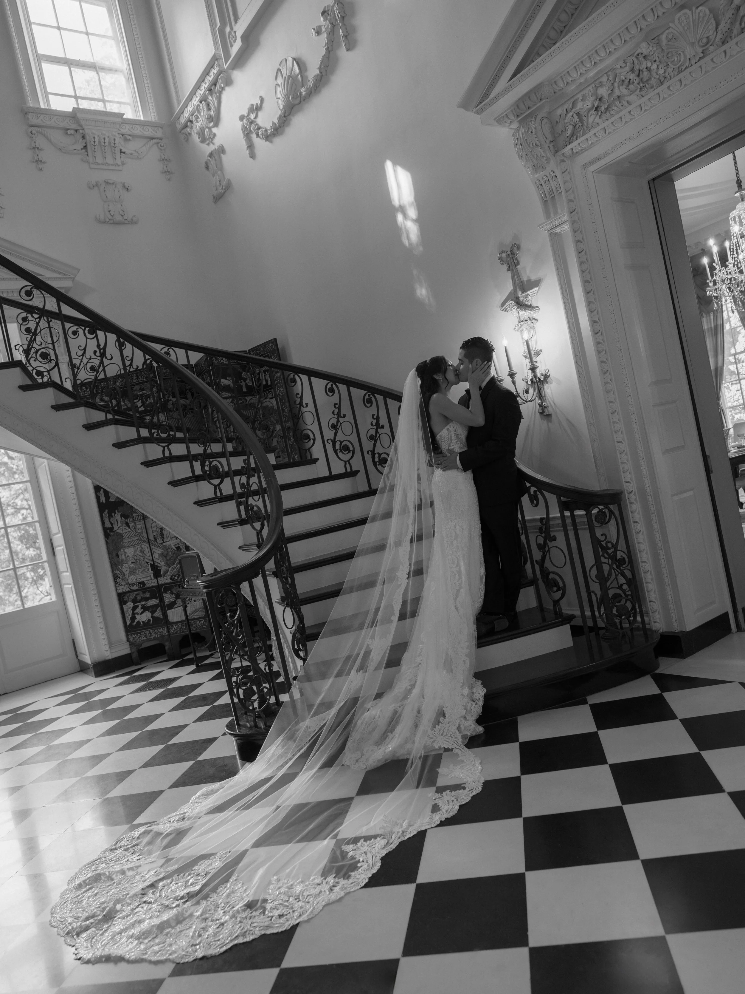 A bride and groom share a kiss on a curved staircase inside an elegant, ornately decorated building with a checkered floor.