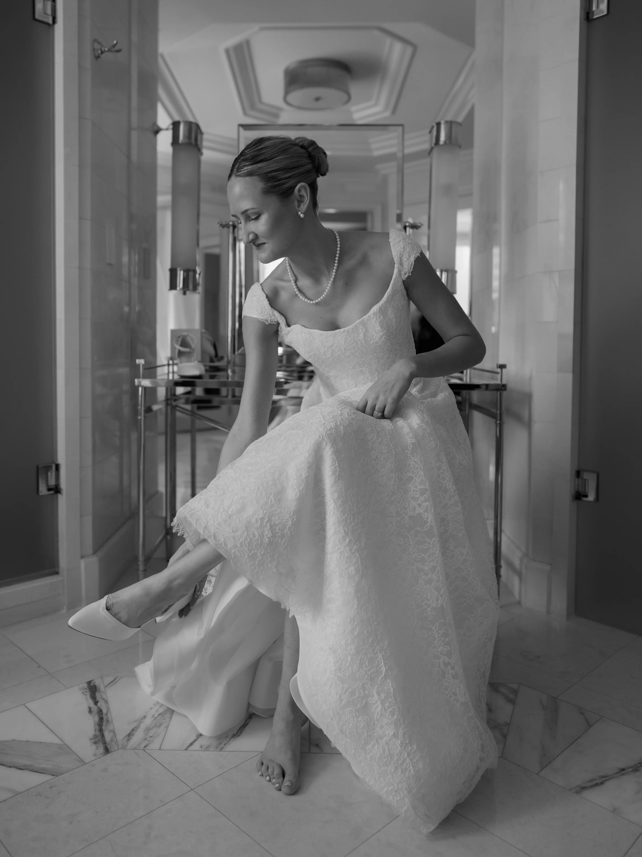 A bride in a wedding dress sitting and adjusting her shoe in a bathroom with marble floors, holding her gown up.