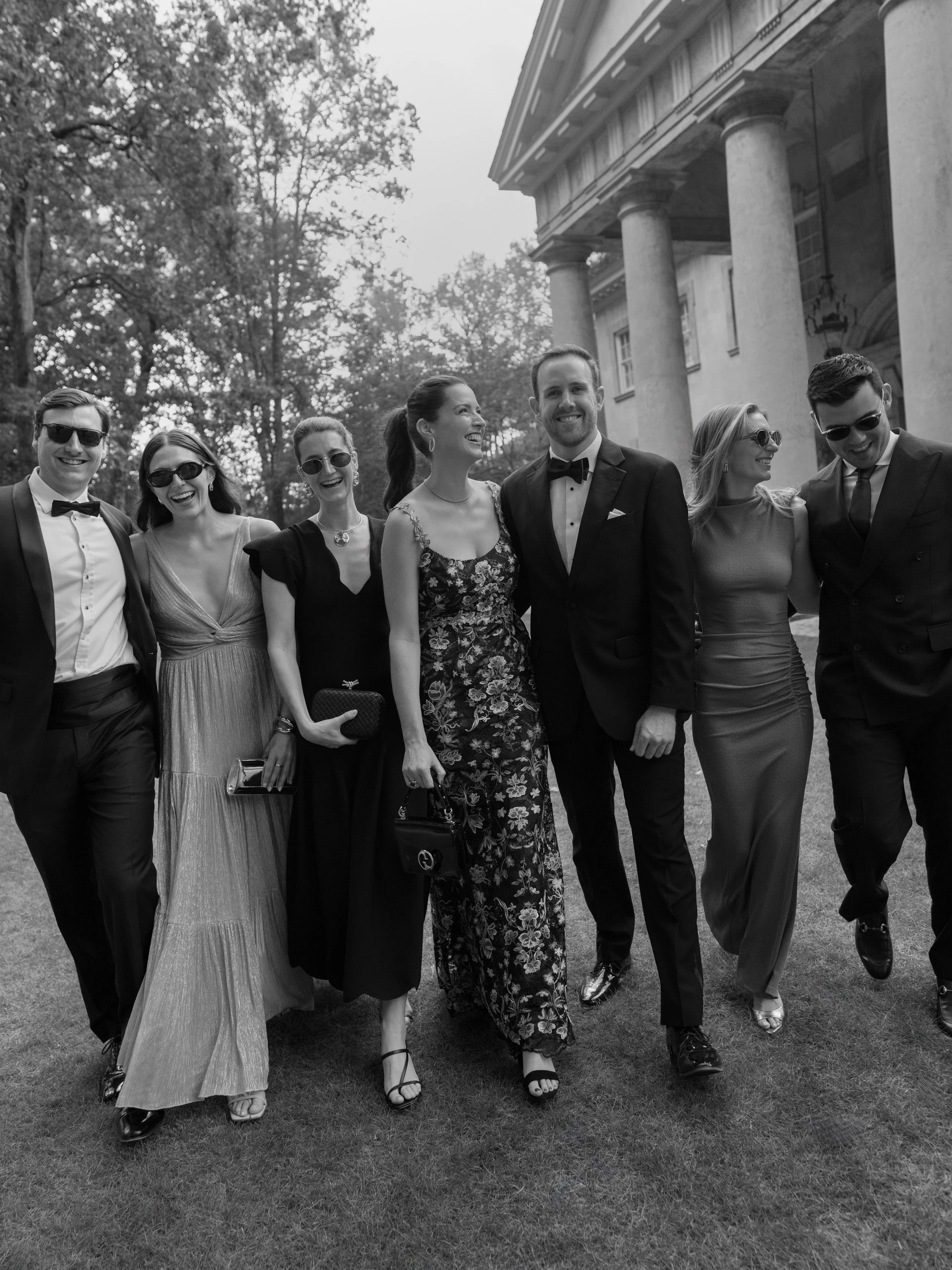 A group of people dressed in formal attire, smiling and walking together in front of a historic building with columns, trees, and grass.