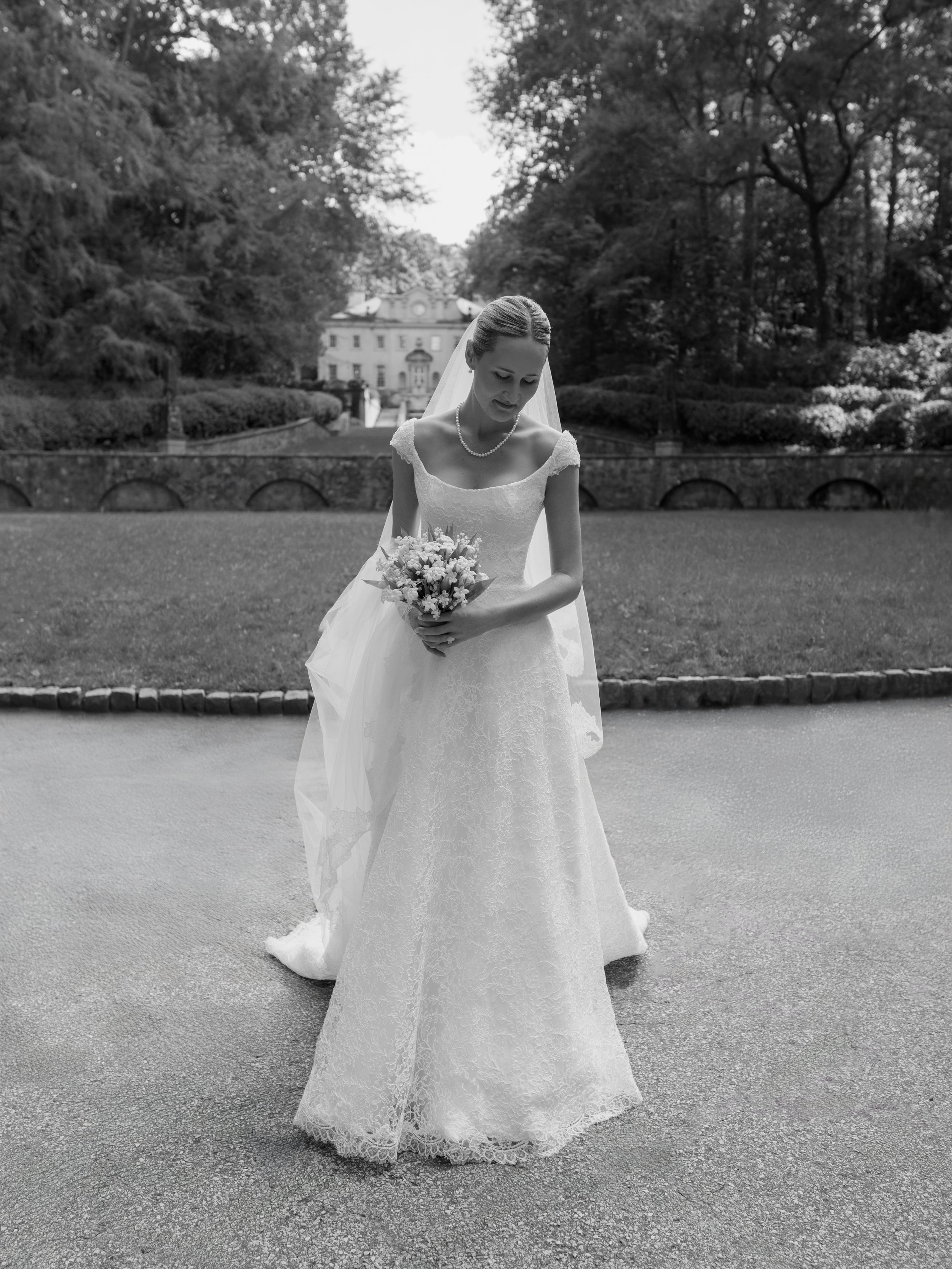 A bride in a lace wedding gown and veil stands outdoors on a paved path, holding a flower bouquet, with trees and a large building in the background.