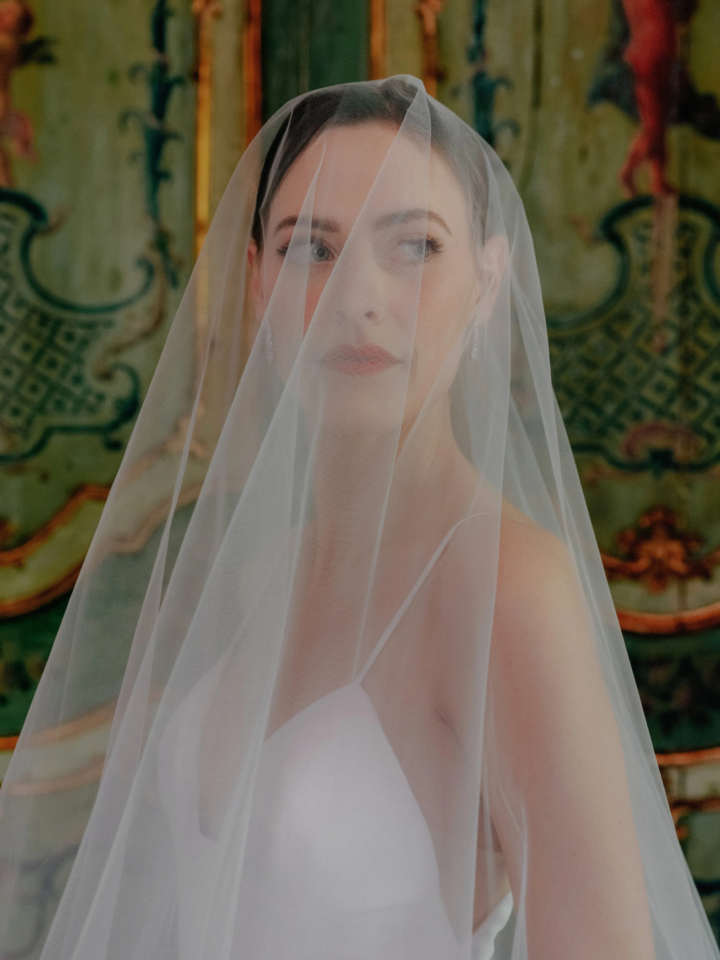 A bride with dark hair and makeup, wearing a white dress and veil, looks to the side in front of an ornate, colorful background.