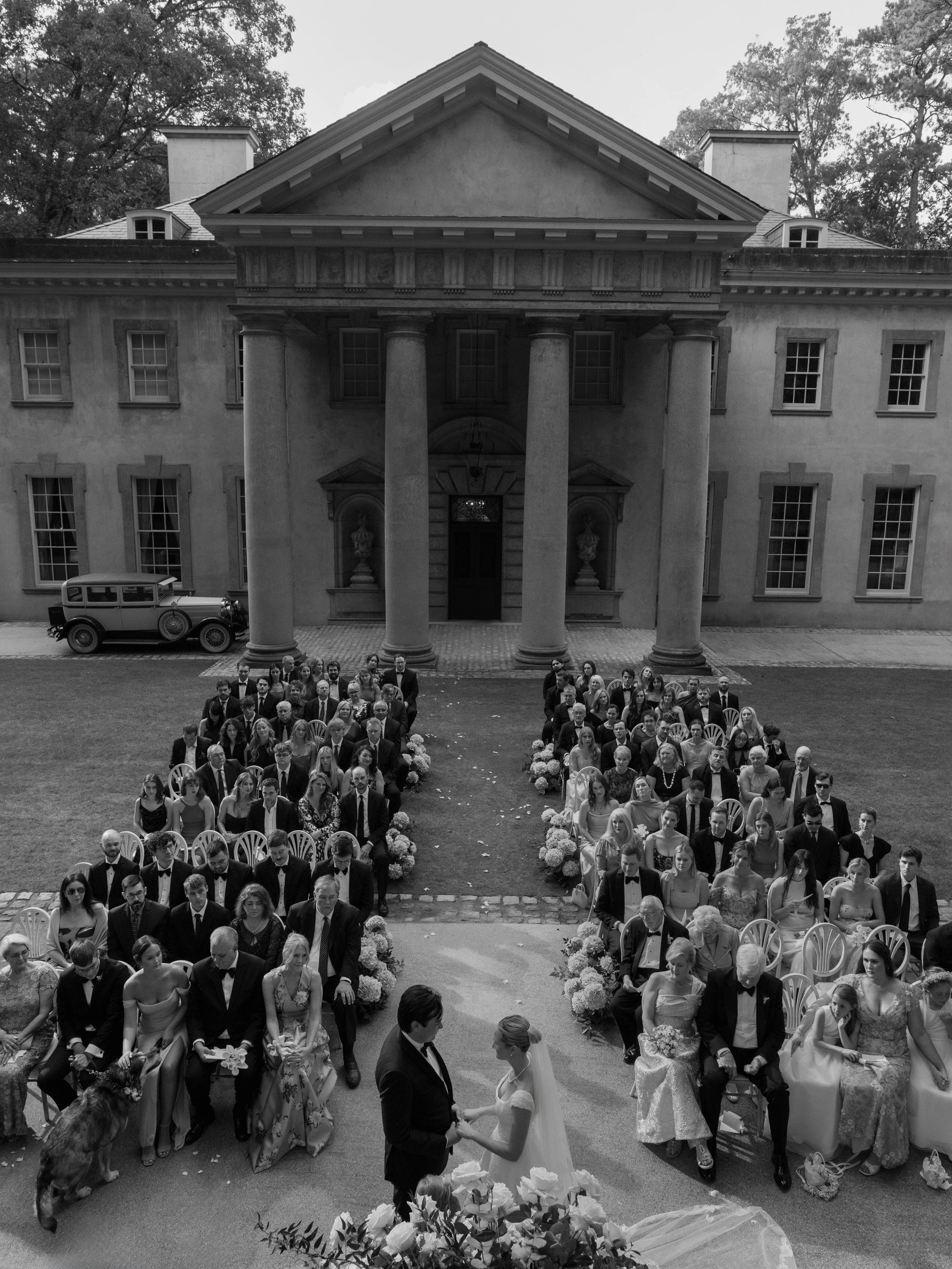A black and white photo of a wedding ceremony outdoors in front of a large classical-style mansion with columns. The bride and groom stand facing each other at the altar, surrounded by seated guests in formal attire, with floral decorations along the
