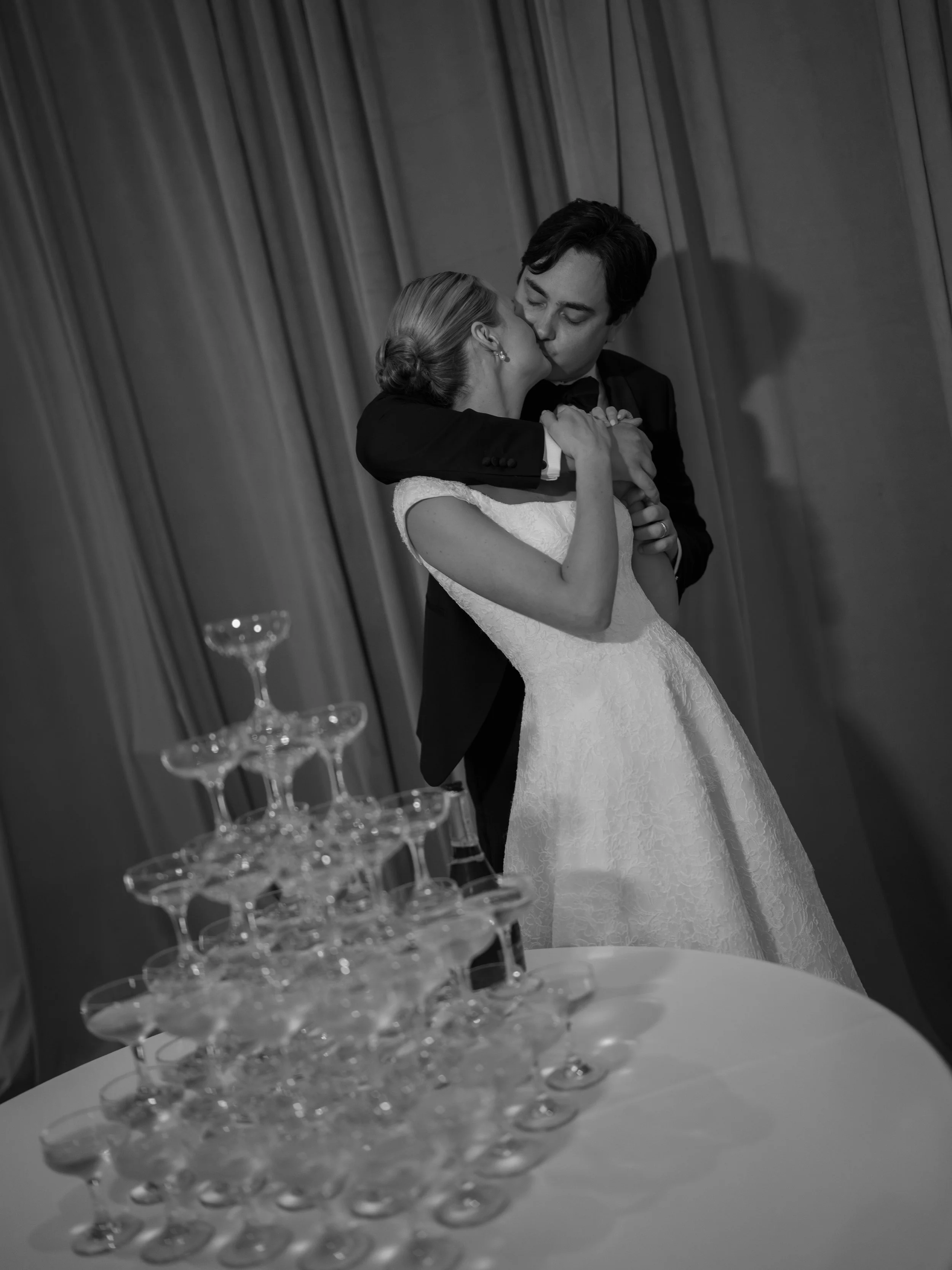 A black and white photo of a couple in wedding attire kissing at a wedding reception. A champagne tower is visible in the foreground, with glasses stacked in a pyramid shape in front of a curtain backdrop.