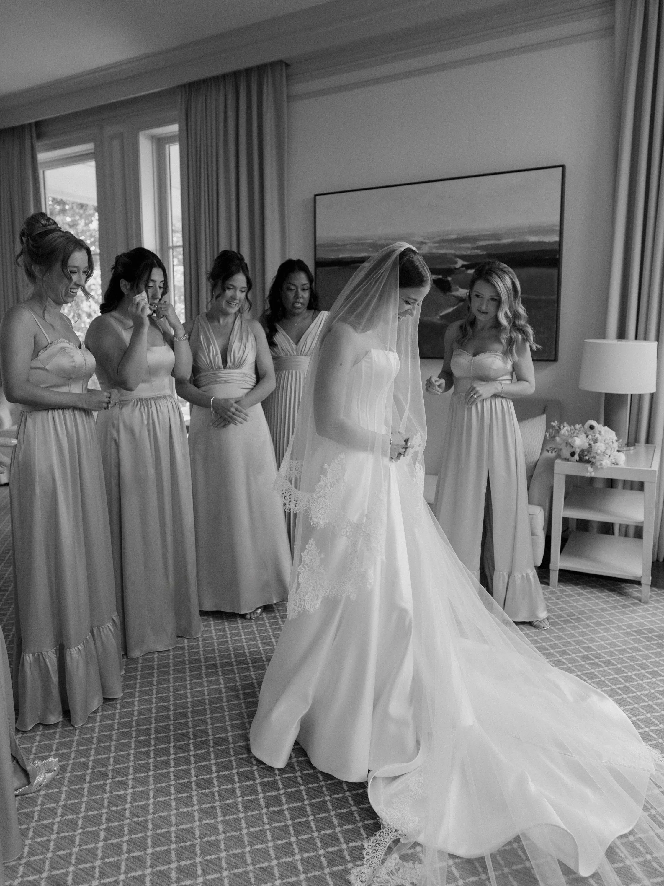 A bride in a wedding dress with a veil, surrounded by her bridesmaids, standing in a hotel room with large windows, curtains, and a landscape painting on the wall.