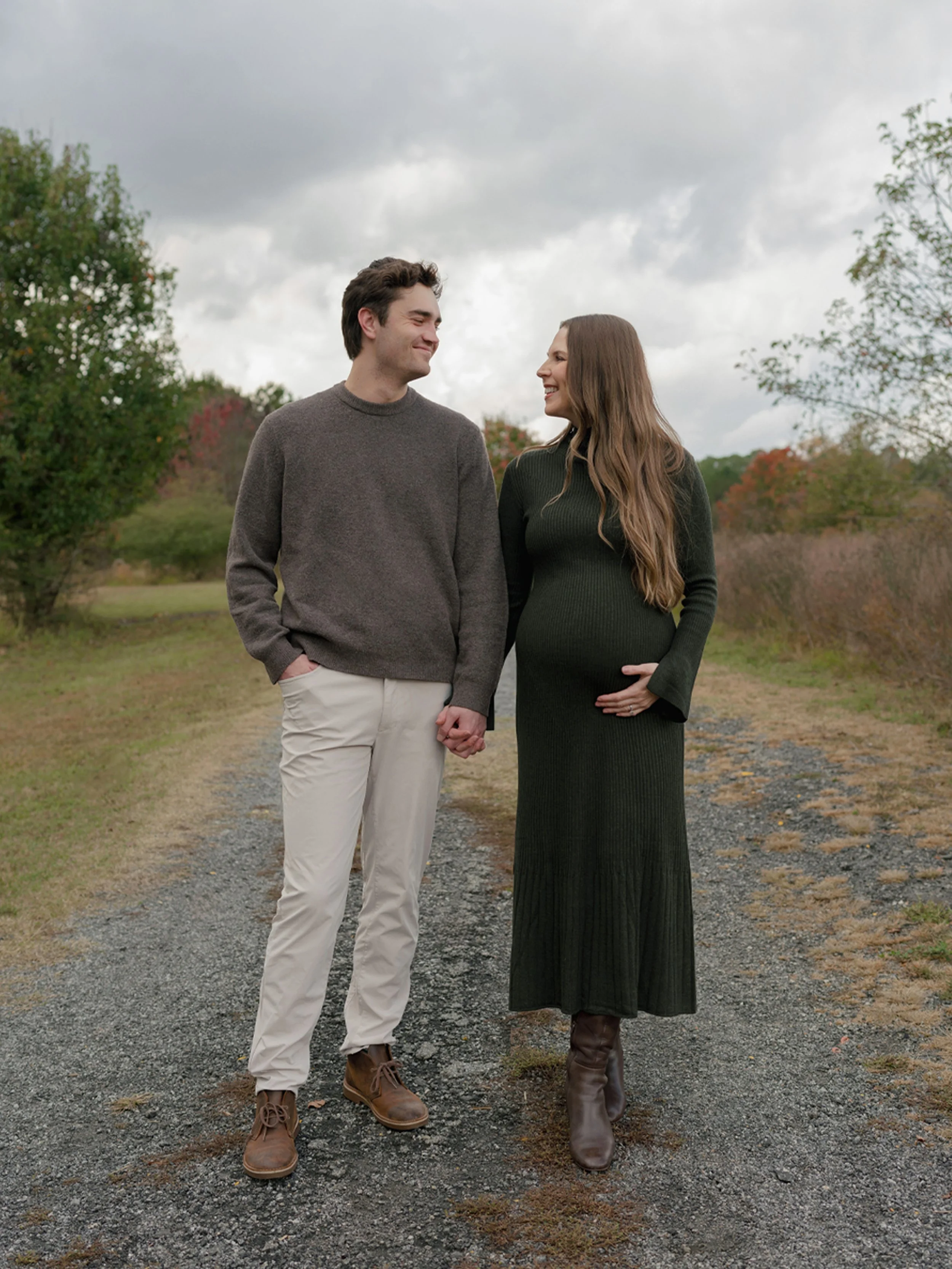 A loving couple holding hands and smiling at each other outdoors on a cloudy day, surrounded by trees with fall foliage.
