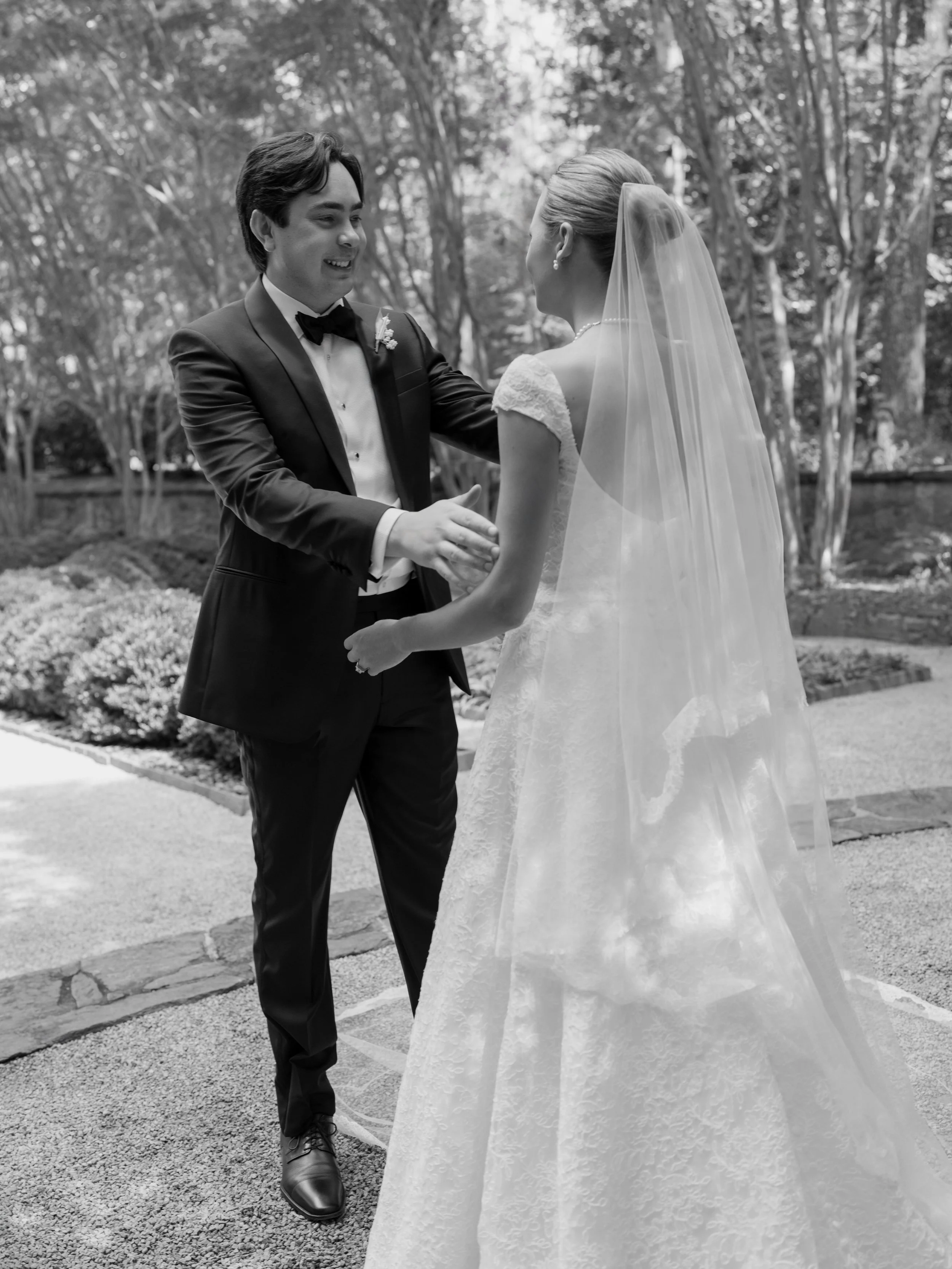 A bride and groom hold hands and smile at each other outdoors in a garden or park setting, dressed in wedding attire with trees in the background.