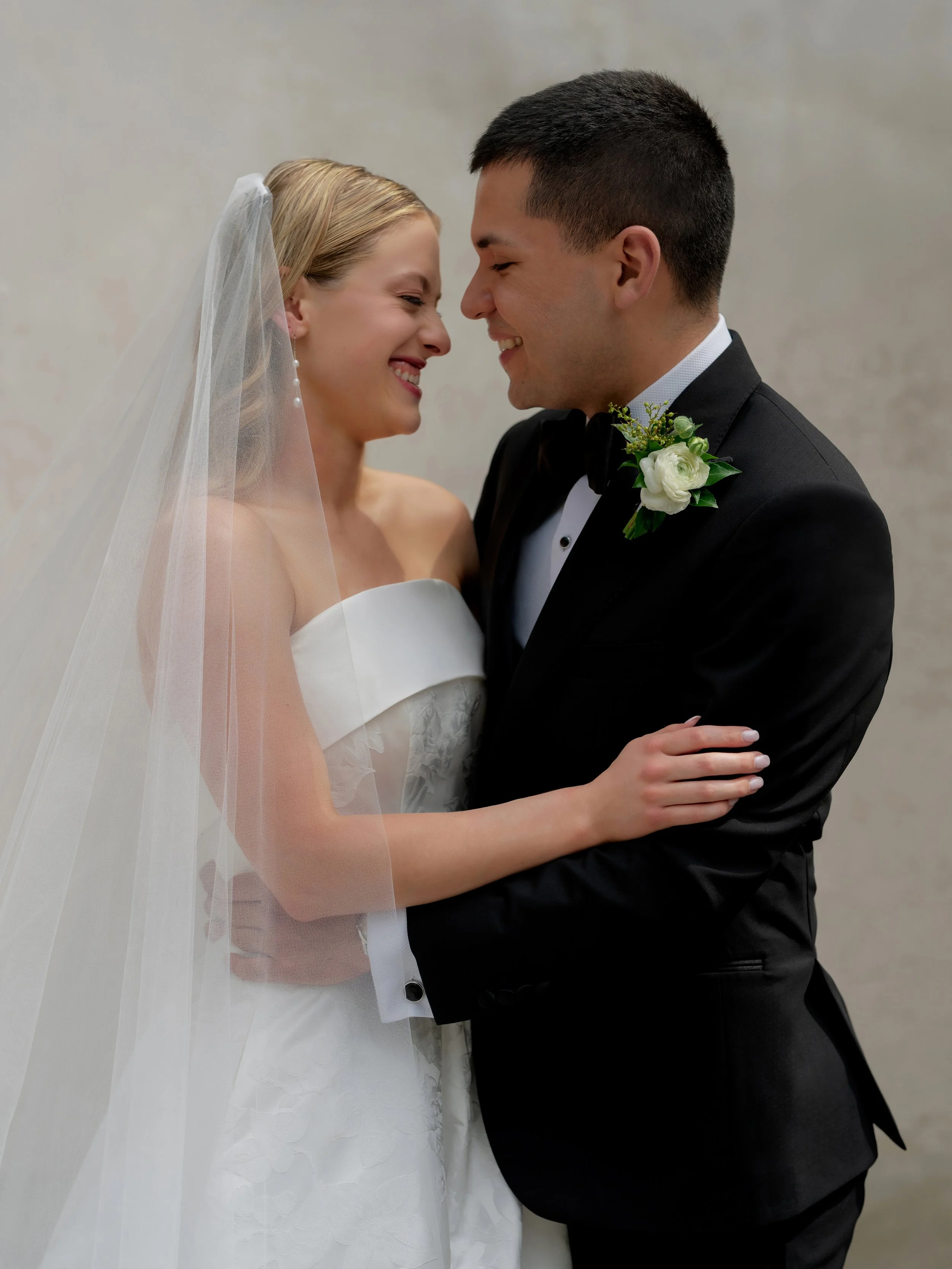 Bride and groom smiling and embracing at their wedding, standing close with foreheads touching, bride wearing a white strapless dress and veil, groom in a black tuxedo with a boutonnière.