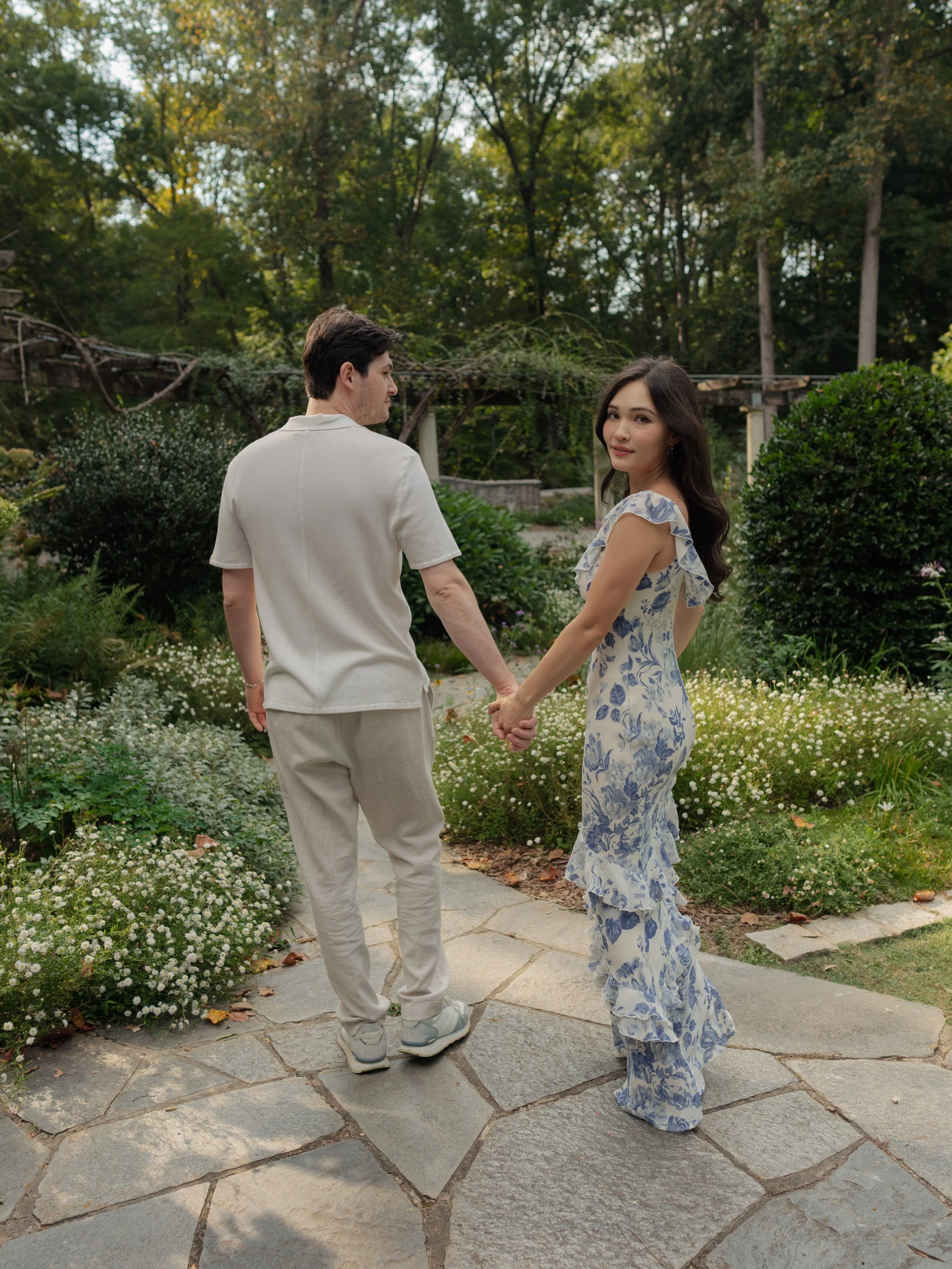 A man and woman holding hands in a garden, surrounded by greenery and small white flowers.