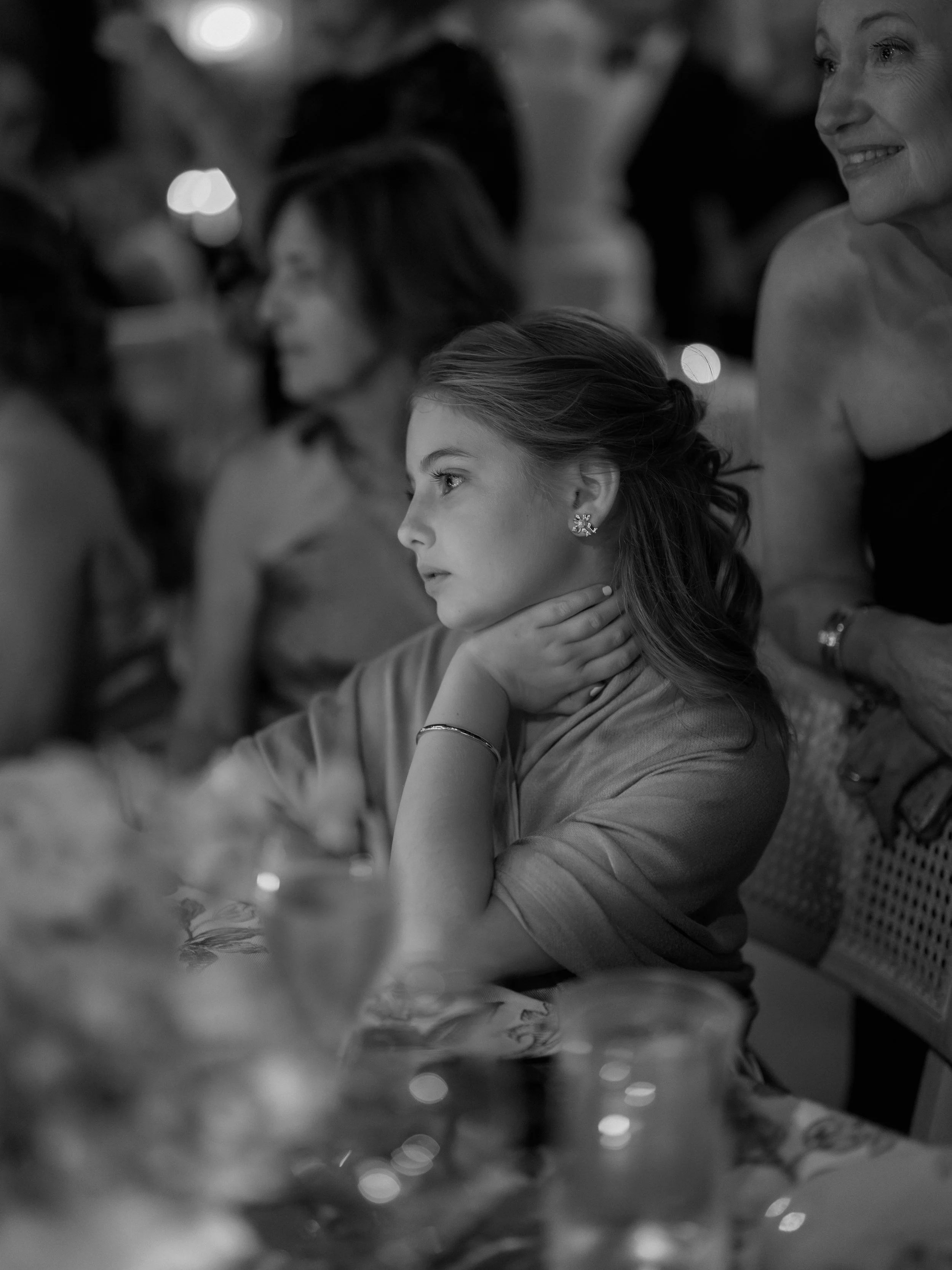A young woman with earrings and a bracelet sitting at a table, looking to the side with her hand around her neck, surrounded by other people at a social event.