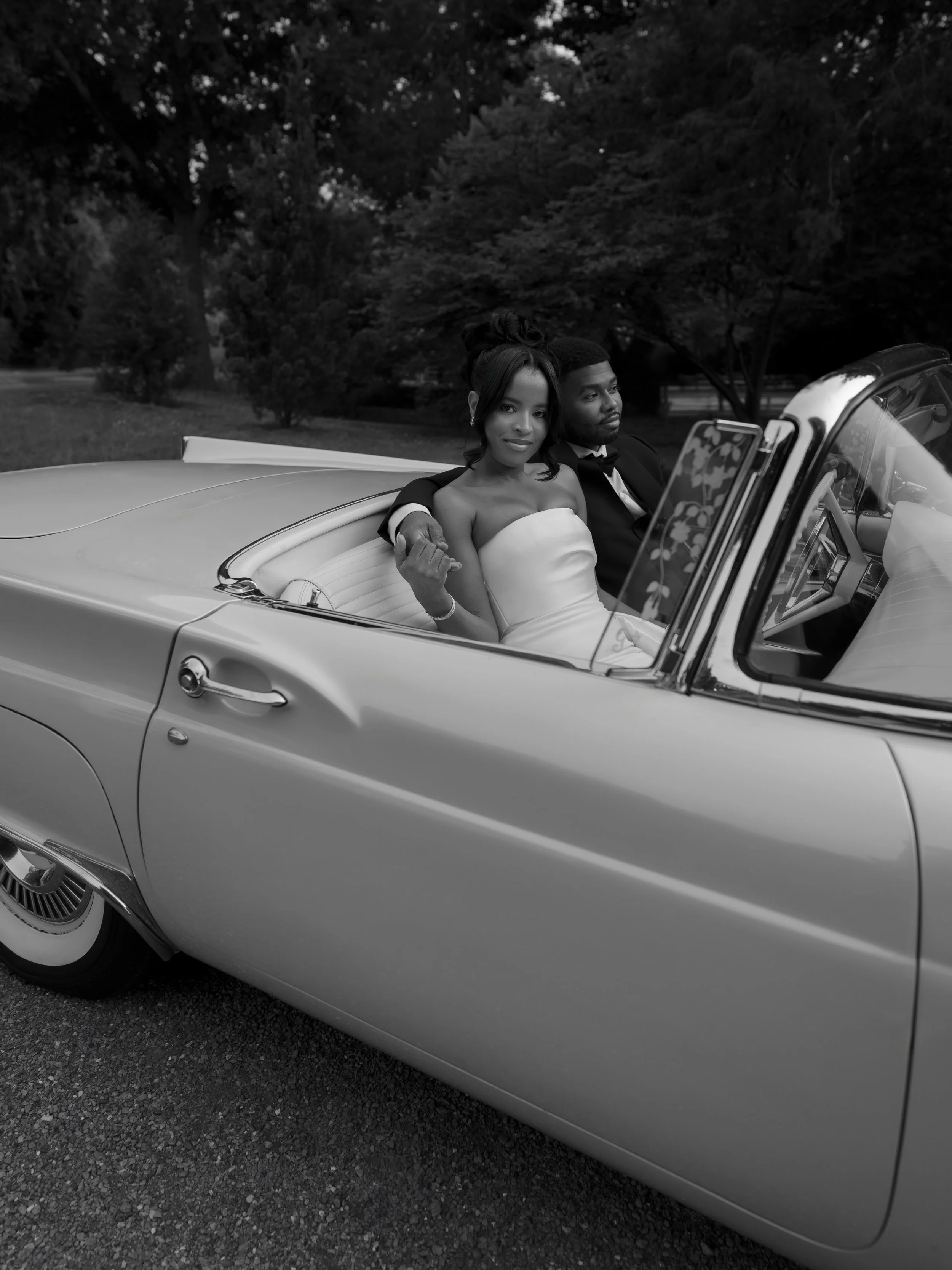 A bride and groom sitting in a vintage convertible car, with trees in the background, the bride wearing a strapless dress and the groom in a tuxedo.