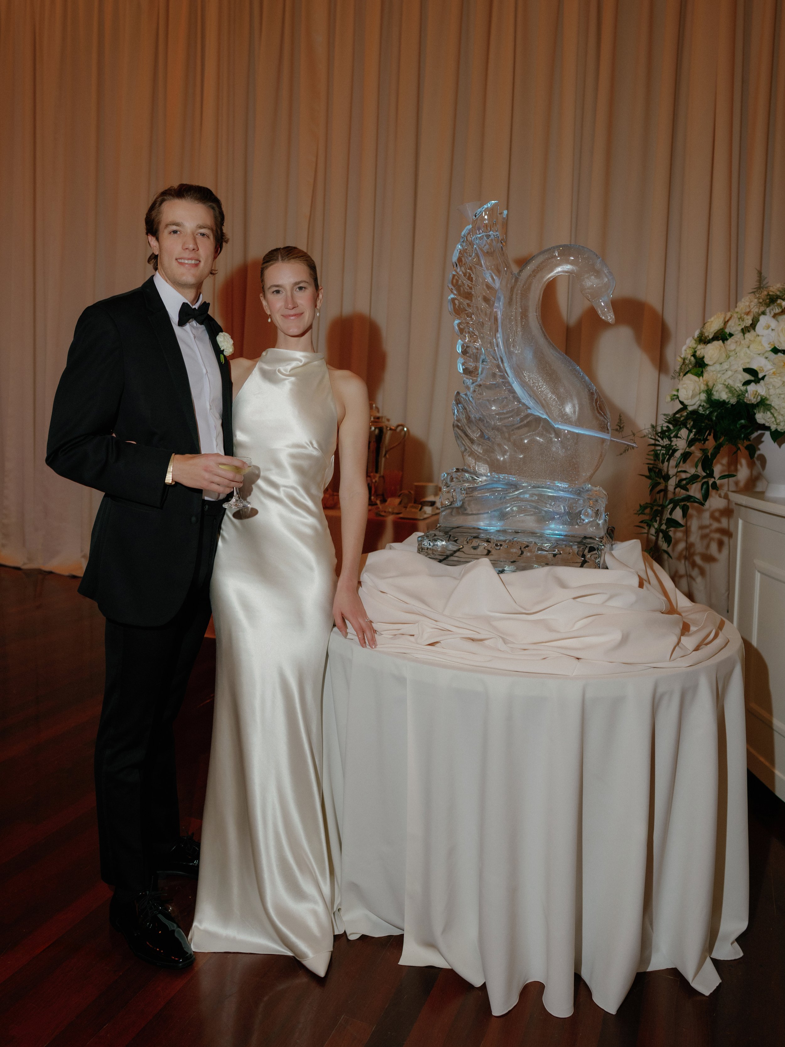 A bride and groom standing together at their wedding reception, dressed in formal attire, beside an ice sculpture of a swan on a table with a white tablecloth, with a floral arrangement in the background.