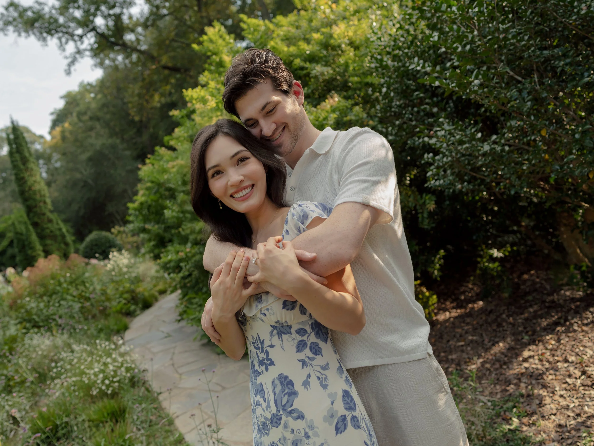 A happy couple embracing outdoors in a lush garden, smiling at each other, with trees and flowering plants in the background.
