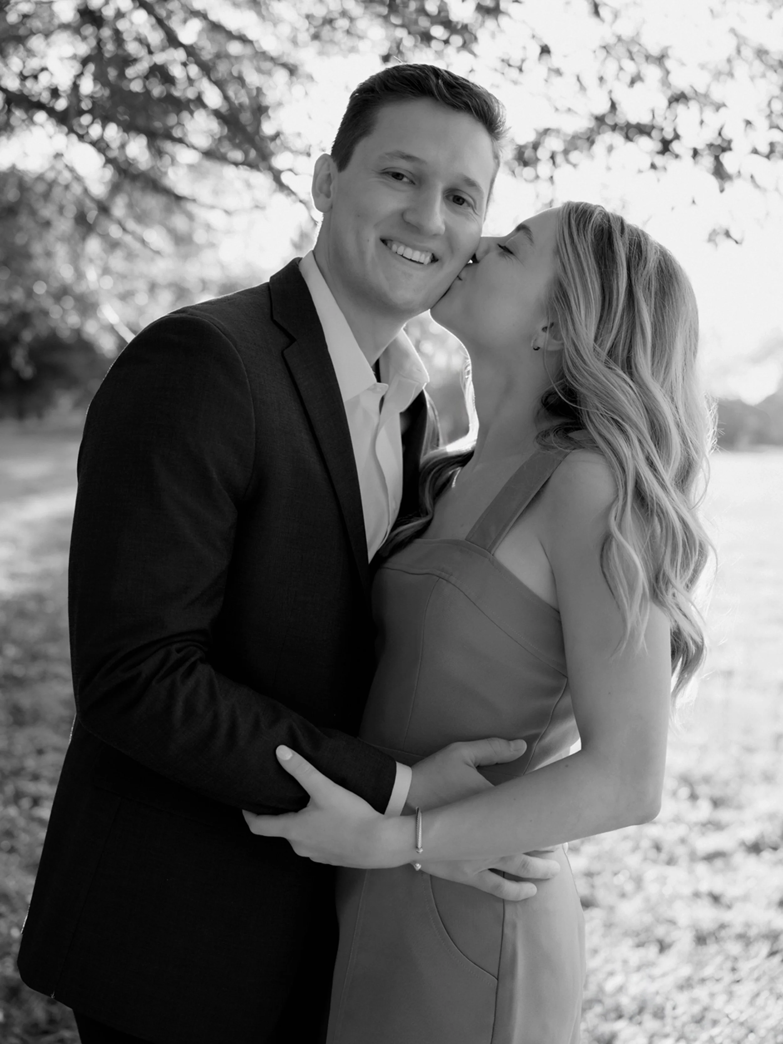 A young couple in formal attire, the woman in a sleeveless dress and the man in a suit, sharing a kiss on the cheek while embracing outdoors with trees and sunlight in the background.