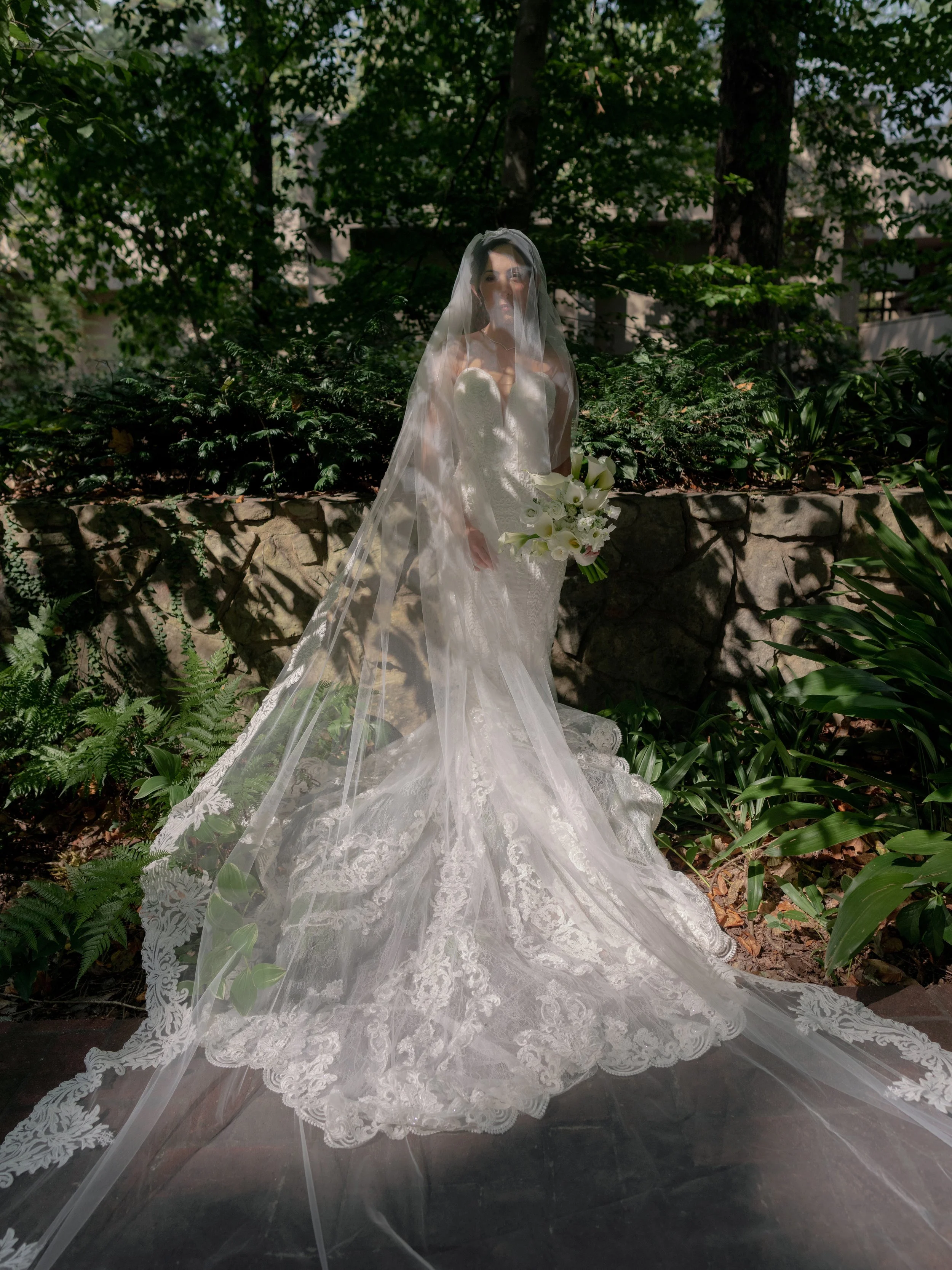 A bride in a strapless wedding gown with lace details and a long veil holding a bouquet of white flowers, standing outdoors in a lush green setting.