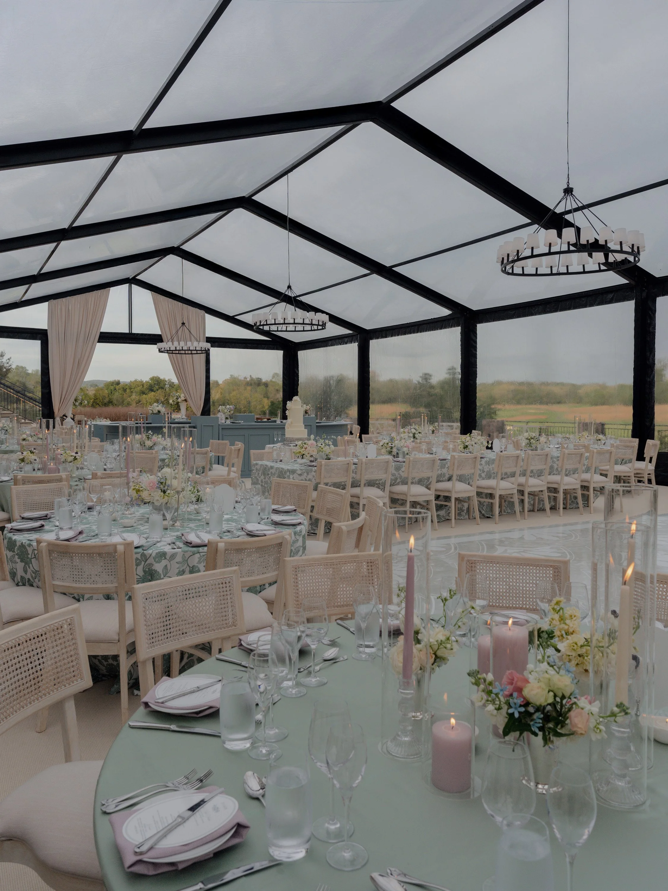 Elegant outdoor wedding reception under a glass tent with round and rectangular tables decorated with pastel flowers and candles, surrounded by white chairs, overlooking a scenic landscape.