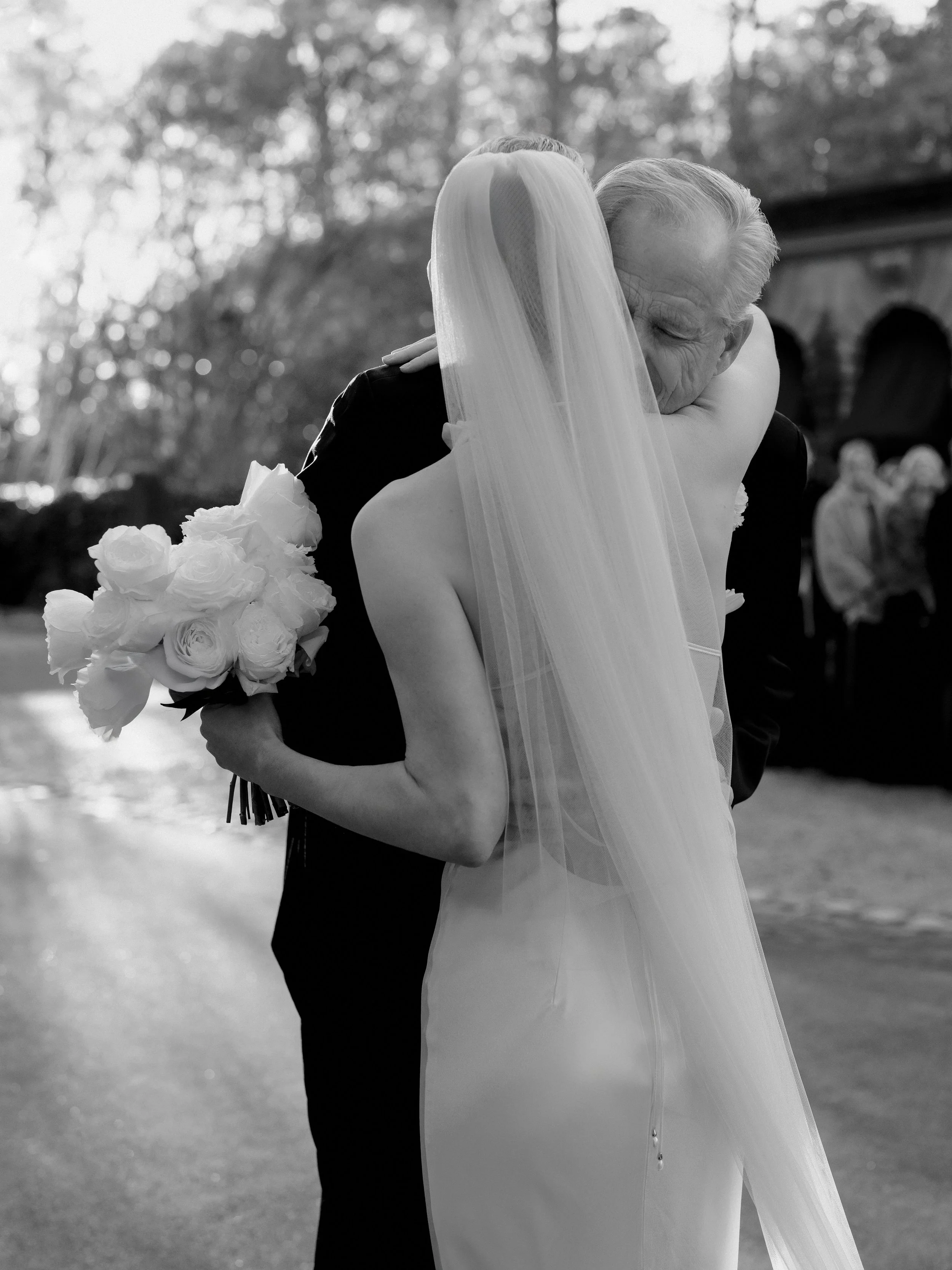 A bride in a white wedding dress and veil hugging an elderly man, who appears emotional, outdoors during the daytime.