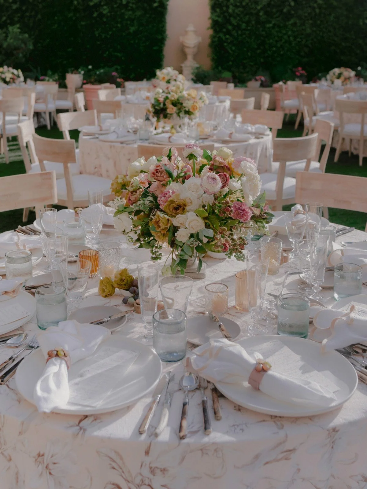 Elegant outdoor wedding reception setup with round tables adorned with floral centerpieces, white tablecloths, and place settings including napkins and glassware.