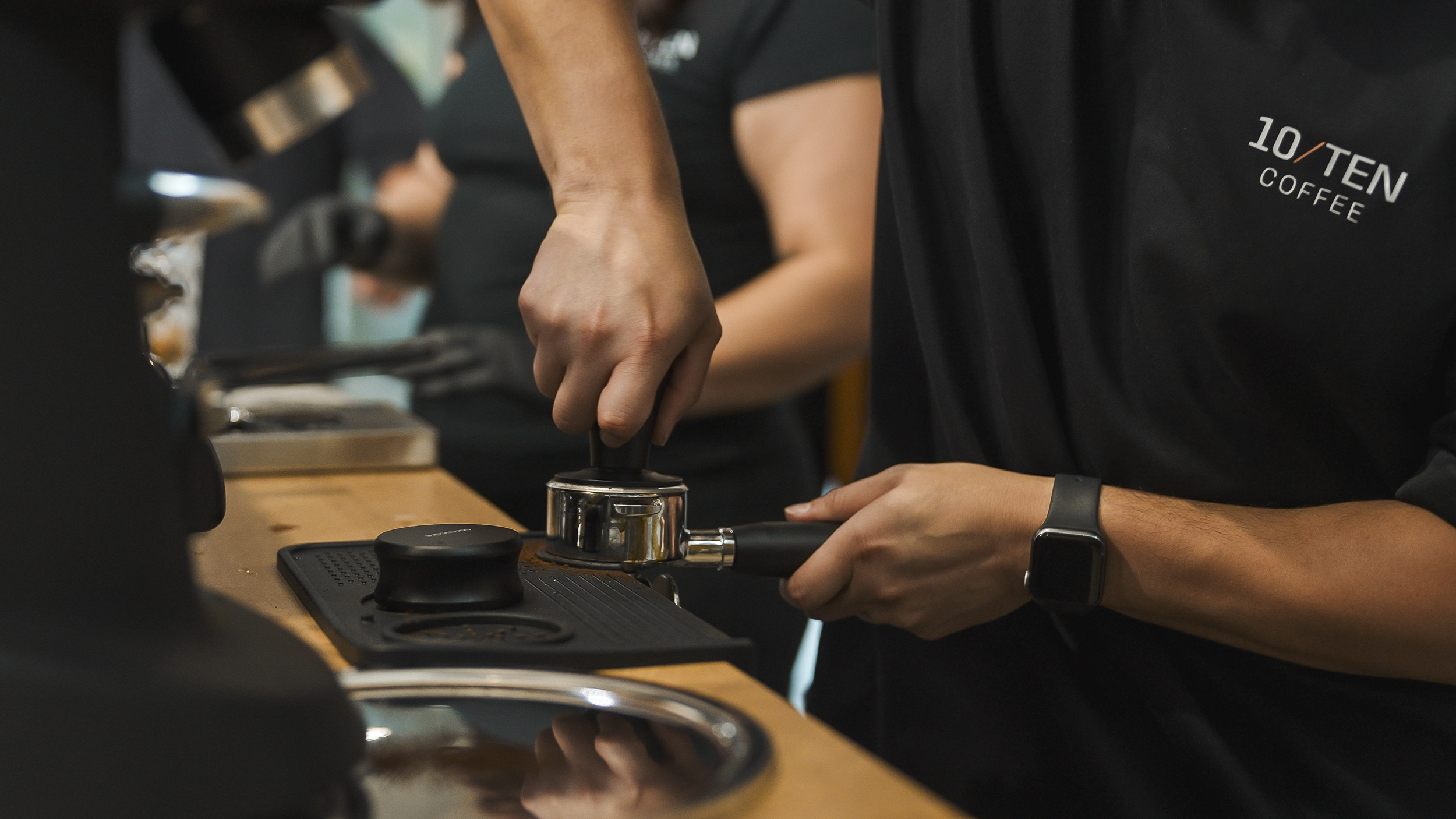 Barista tamping coffee grounds in a metal portafilter at a coffee shop counter.