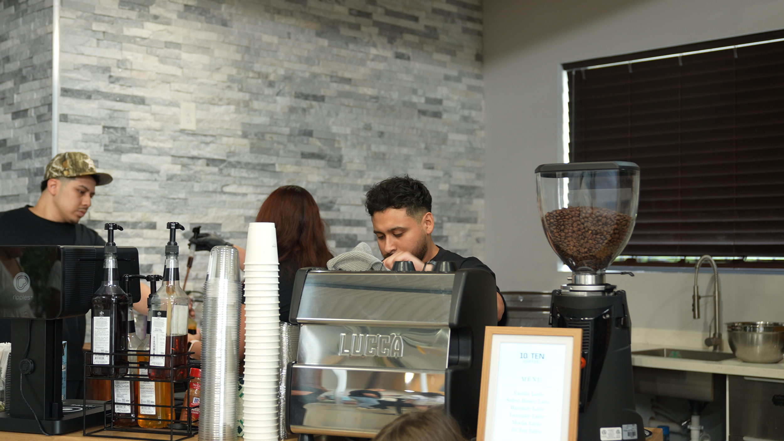 Barista preparing coffee behind the counter at a coffee shop with a coffee grinder filled with coffee beans on the side.
