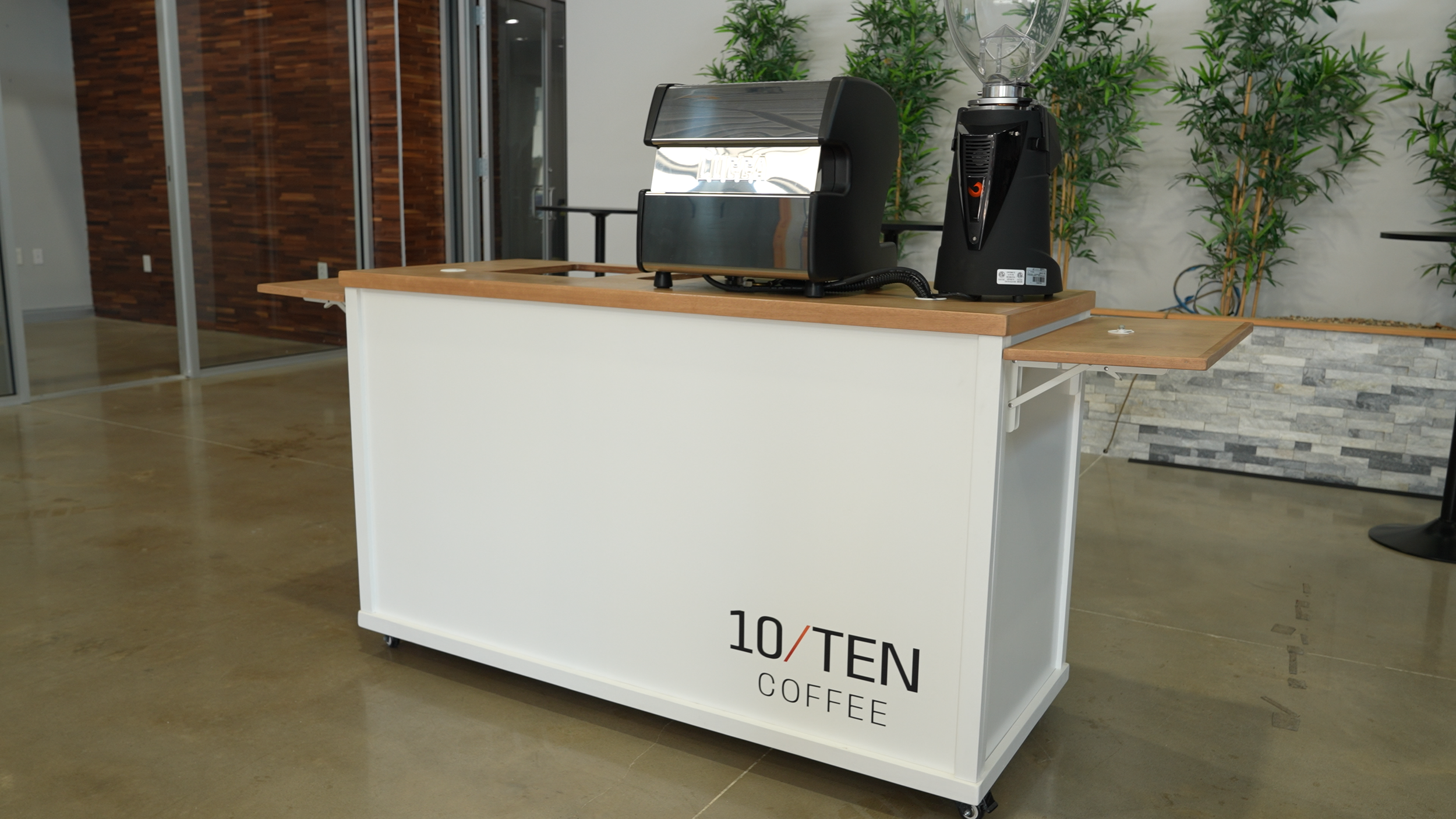 A coffee station with a coffee grinder and a commercial coffee machine on a white mobile counter, in a modern indoor setting with wooden and stone wall accents and green plants in the background.