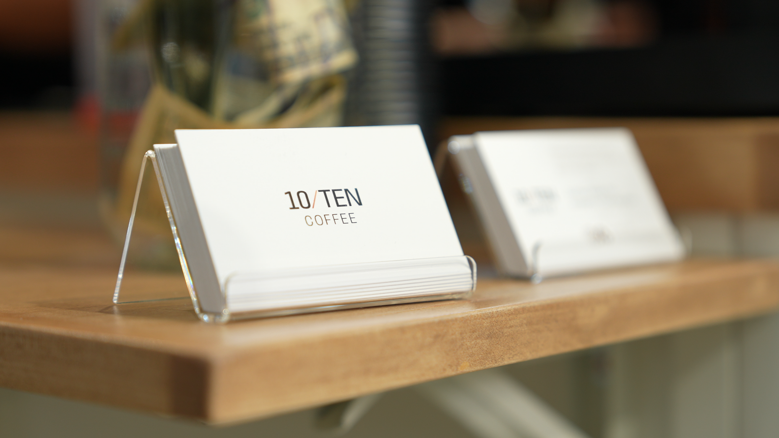 Close-up of a white card display on a wooden surface with the text '10/TEN COFFEE' printed on it, blurred background.