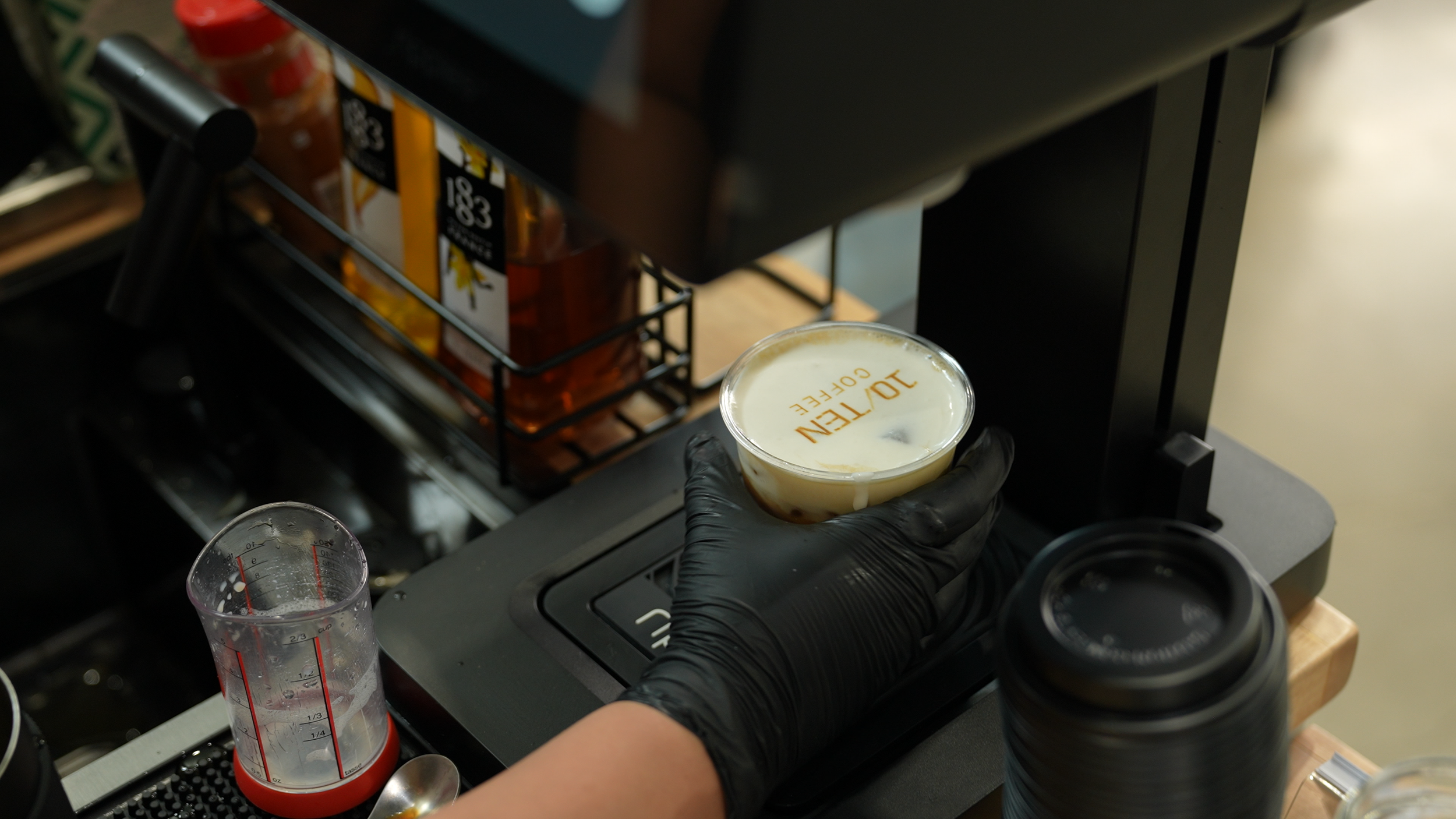 A person wearing black gloves holding a cup of coffee with the word 'JOTTEEN COFFEE' on the surface, in a coffee shop or café setting with coffee brewing equipment and a measuring cup nearby.