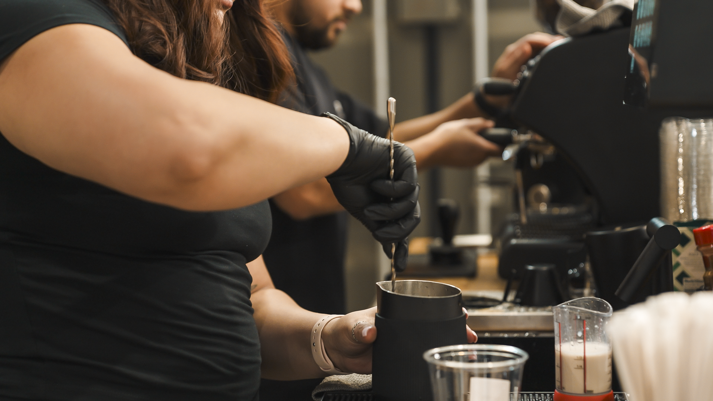 Two people preparing beverages in a cafe; one is holding a metal cup while the other is operating an espresso machine, surrounded by coffee-making tools and ingredients.